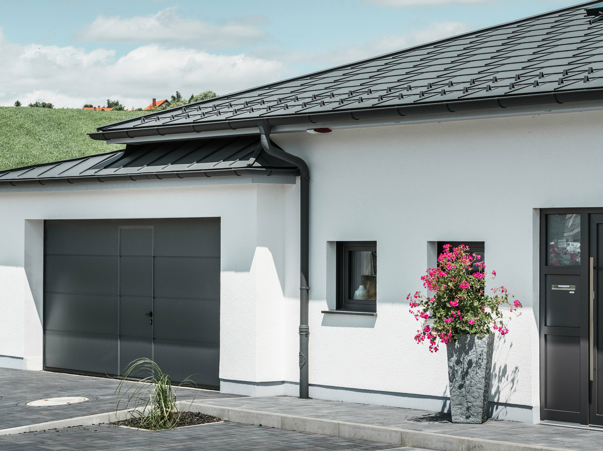 Das Bild präsentiert die Vorderseite eines Einfamilienhauses in Ilmenau, Thüringen, mit einem eleganten PREFA Dach in P.10 Dunkelgrau. Die Dachplatte R.16 verleiht dem Haus ein modernes Aussehen, während das integrierte PREFA Dachentwässerungssystem mit seiner dunkelgrauen Hängerinne und dem Ablaufrohr effizienten Abfluss von Regenwasser garantieren. Die Garage, ausgestattet mit dunkelgrauem PREFALZ, fügt sich harmonisch in die Gesamtästhetik ein und unterstreicht die Qualität und den Stil von PREFA Aluminiumprodukten.