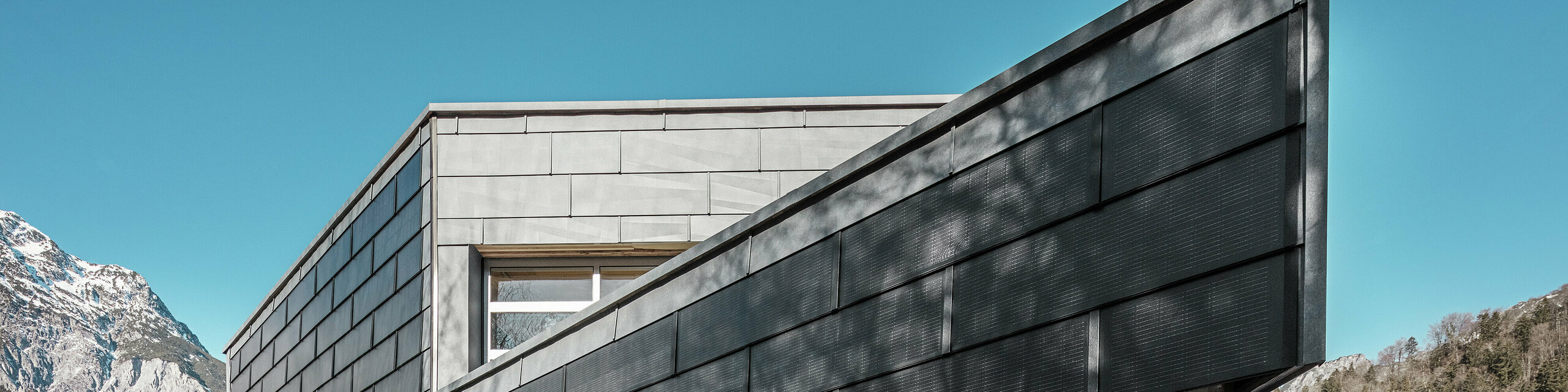 Das Firmengebäude der Spenglerei RÜMA in Vorarlberg beeindruckt mit seiner modernen Fassade aus PREFA Solardachplatten und Sichtbeton. Umgeben von Bergen und Bäumen, deren Schatten sich auf der Wand abzeichnen, steht es unter einem klaren, blauen Himmel. Die spitze Gebäudeecke und die PREFA Fassadenpaneele FX.12 betonen das markante Erscheinungsbild zusätzlich.