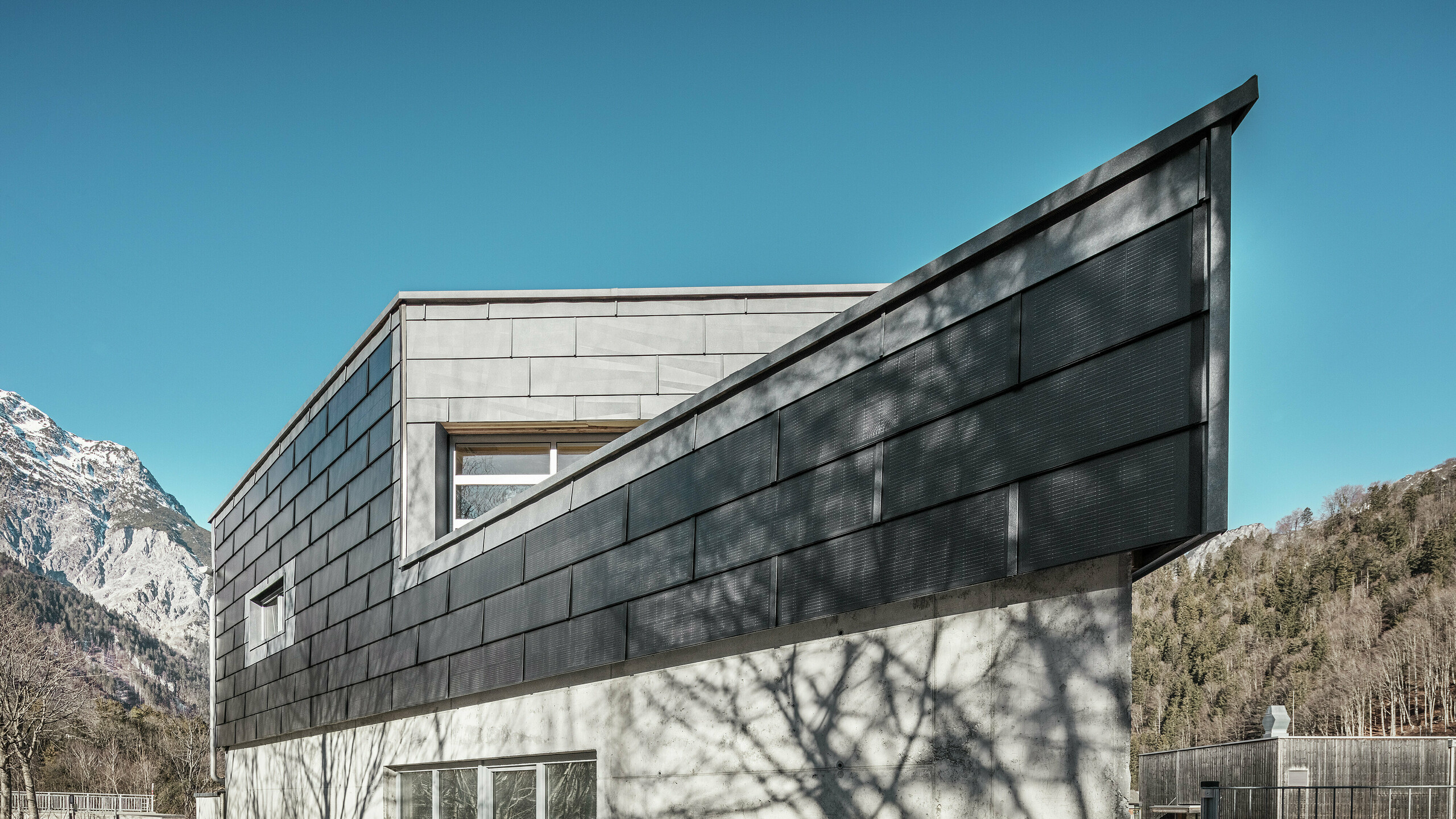 Das Firmengebäude der Spenglerei RÜMA in Vorarlberg beeindruckt mit seiner modernen Fassade aus PREFA Solardachplatten und Sichtbeton. Umgeben von Bergen und Bäumen, deren Schatten sich auf der Wand abzeichnen, steht es unter einem klaren, blauen Himmel. Die spitze Gebäudeecke und die PREFA Fassadenpaneele FX.12 betonen das markante Erscheinungsbild zusätzlich.