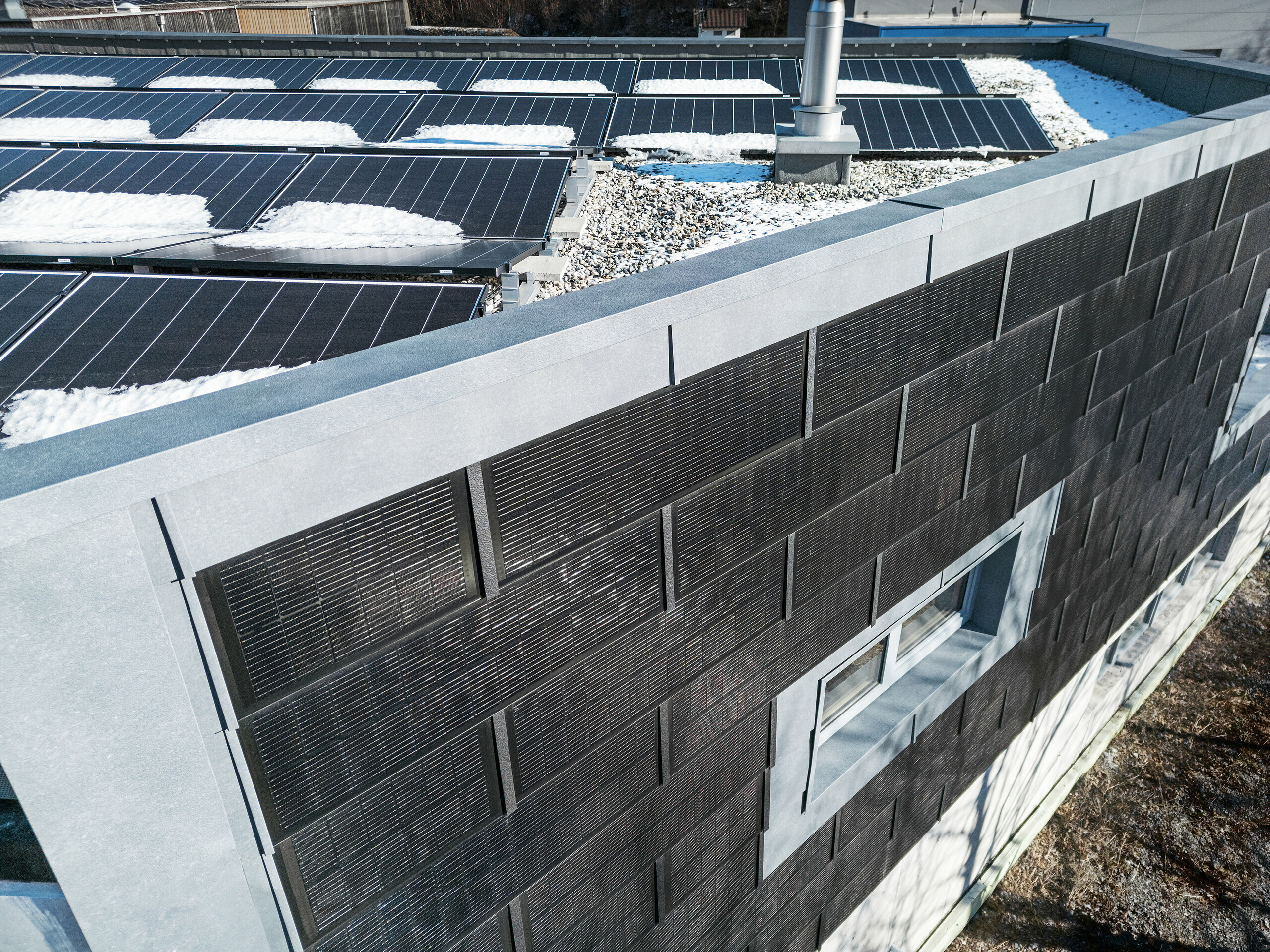 Die Draufsicht zeigt die Fassade der Spenglerei RÜMA in Vorarlberg mit grauen Fassadenpaneelen FX.12 sowie innovativen PREFA Solardachplatten mit integrierten Photovoltaikmodulen. Im Vordergrund ist das teilweise mit Schnee bedeckte Flachdach mit weiteren Photovoltaikmodulen zu sehen.