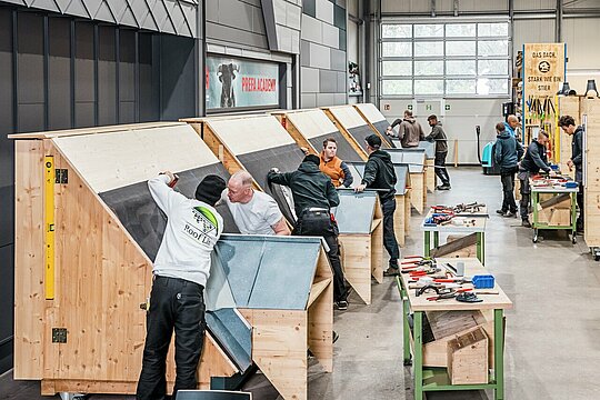In einer modernen Werkhalle der PREFA Academy arbeiten mehrere Handwerker an speziell konstruierten Holzmodellen mit Dachflächen. Die Teilnehmer üben die fachgerechte Montage von PREFA Aluminiumprodukten. Auf den Arbeitstischen liegen Werkzeuge wie Zangen, Schraubenschlüssel und Messgeräte, die für präzises Arbeiten notwendig sind. Im Hintergrund sind weitere Schulungsstationen und eine große Fensterfront zu sehen, die für helles Tageslicht sorgt. Die Szene vermittelt praxisorientiertes Lernen, bei dem theoretisches Wissen direkt in handwerkliche Fähigkeiten umgesetzt wird, um die Kompetenz und Qualität im Arbeitsalltag zu stärken.