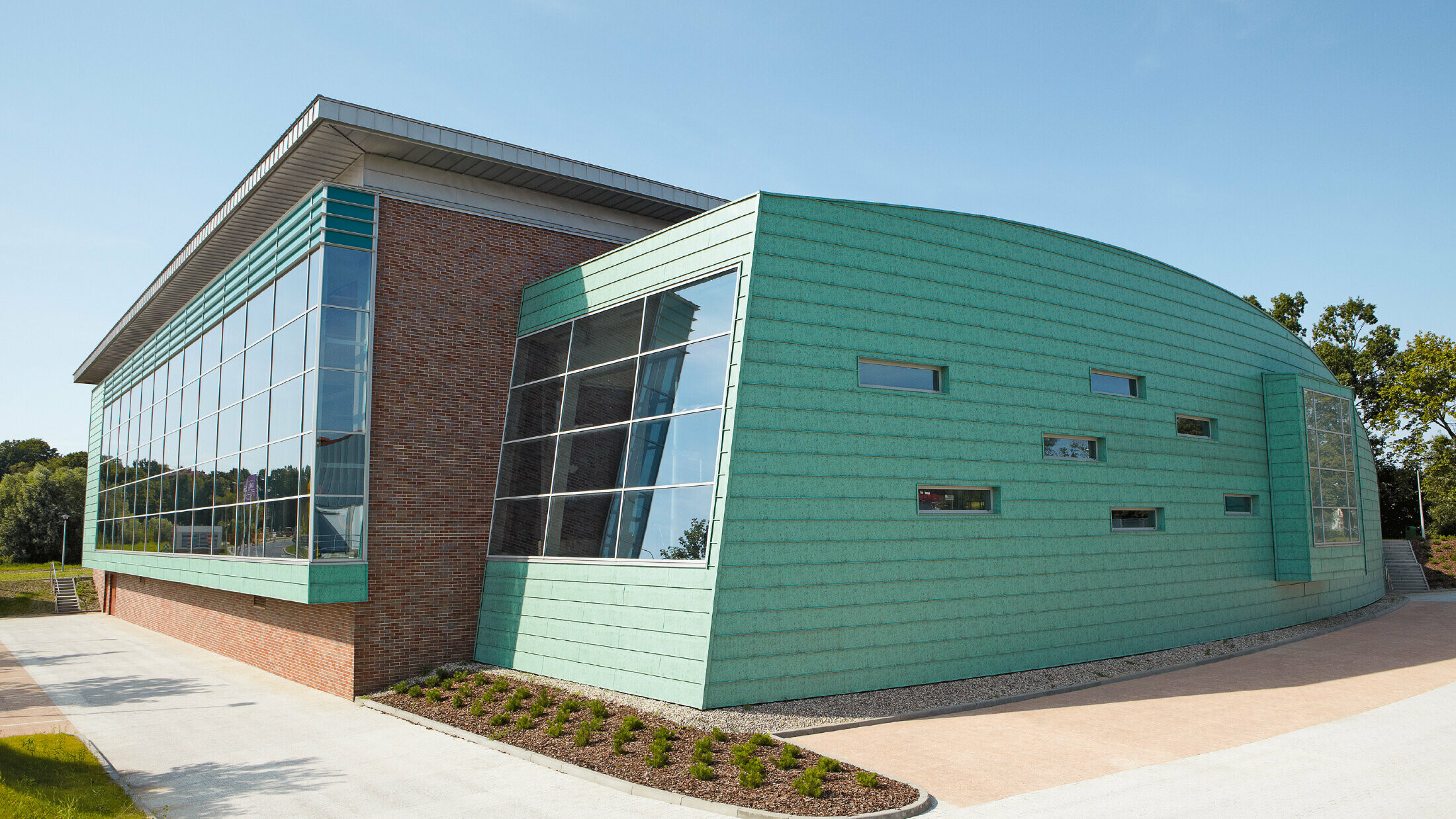 Modernes Schwimmbadgebäude mit auffälliger Fassade aus patinagrünen Aluminium-Paneelen und großen Glasflächen. Der Bau kombiniert gebogene und kantige Formen sowie unterschiedliche Materialien wie Metall und Backstein. Kleine, schmale Fenster lockern die Fassade auf. Das Gebäude steht in einer gepflegten Umgebung unter klarem, blauem Himmel.