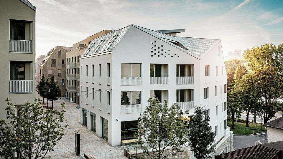 Modernes Wohnquartier Vydrica in Bratislava mit klarer Architektursprache und ruhiger Gassenatmosphäre: Die steil geneigten Satteldächer mit markanten Dachfenstern sind mit PREFA Aluminium Dachschindeln in P.10 Prefaweiß, Bronze und P.10 Sandbraun gedeckt und bilden einen eleganten Eyecatcher im Sonnenlicht. Die präzise ausgeführten Linien und homogenen Flächen unterstreichen die hochwertige Gestaltung. PREFA Aluminium überzeugt durch Langlebigkeit, geringes Gewicht und hohe Witterungsbeständigkeit – ideal für komplexe Dachformen und eine nachhaltige, wartungsarme Gebäudehülle.