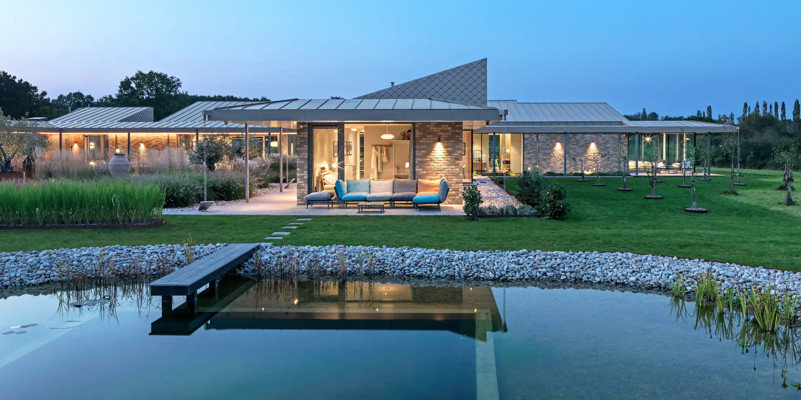 Abendansicht eines traumhaften Wohnhauses in West Sussex mit PREFALZ Aluminiumdach und Fassadenakzenten aus PREFA Wandrauten 20 × 20 in P.10 Bronze. Im Vordergrund ein angelegter Naturpool mit Holzsteg, dahinter eine großzügige Terrasse mit Loungemöbeln und beleuchteter Veranda. Die klare Linienführung der Architektur und die hochwertigen PREFA Produkte schaffen eine elegante Verbindung von Design, Funktionalität und Nachhaltigkeit.