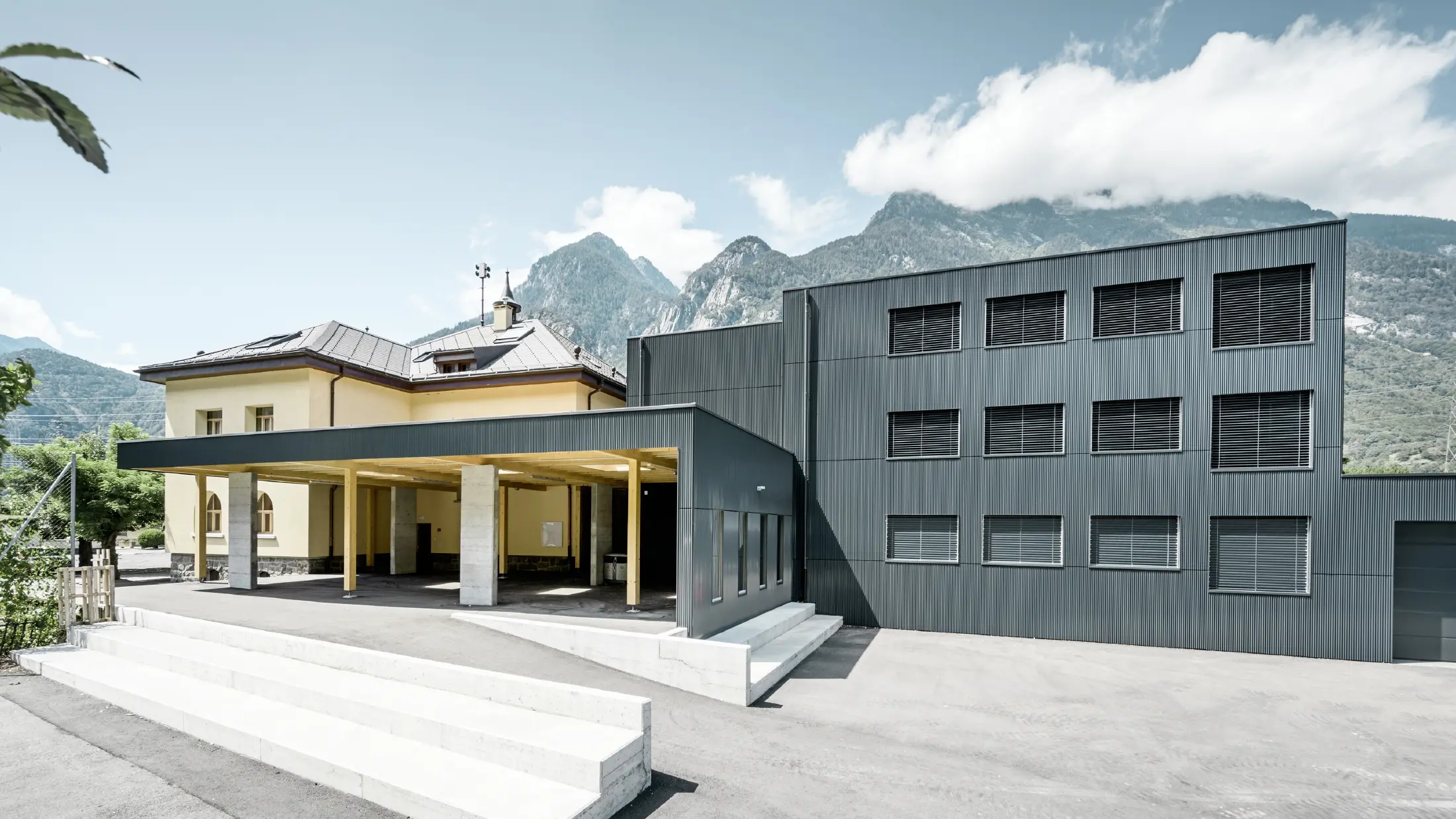 Schulgebäude mit einer Kombination aus historischem, gelb verputztem Altbau und modernem, anthrazitfarbenem Neubau mit Aluminiumfassade im Zackenprofil. Der Eingangsbereich ist überdacht und mit hellen Stützen aus Holz und Beton gestaltet. Im Hintergrund sind imposante Berge und ein teilweise bewölkter Himmel zu sehen.