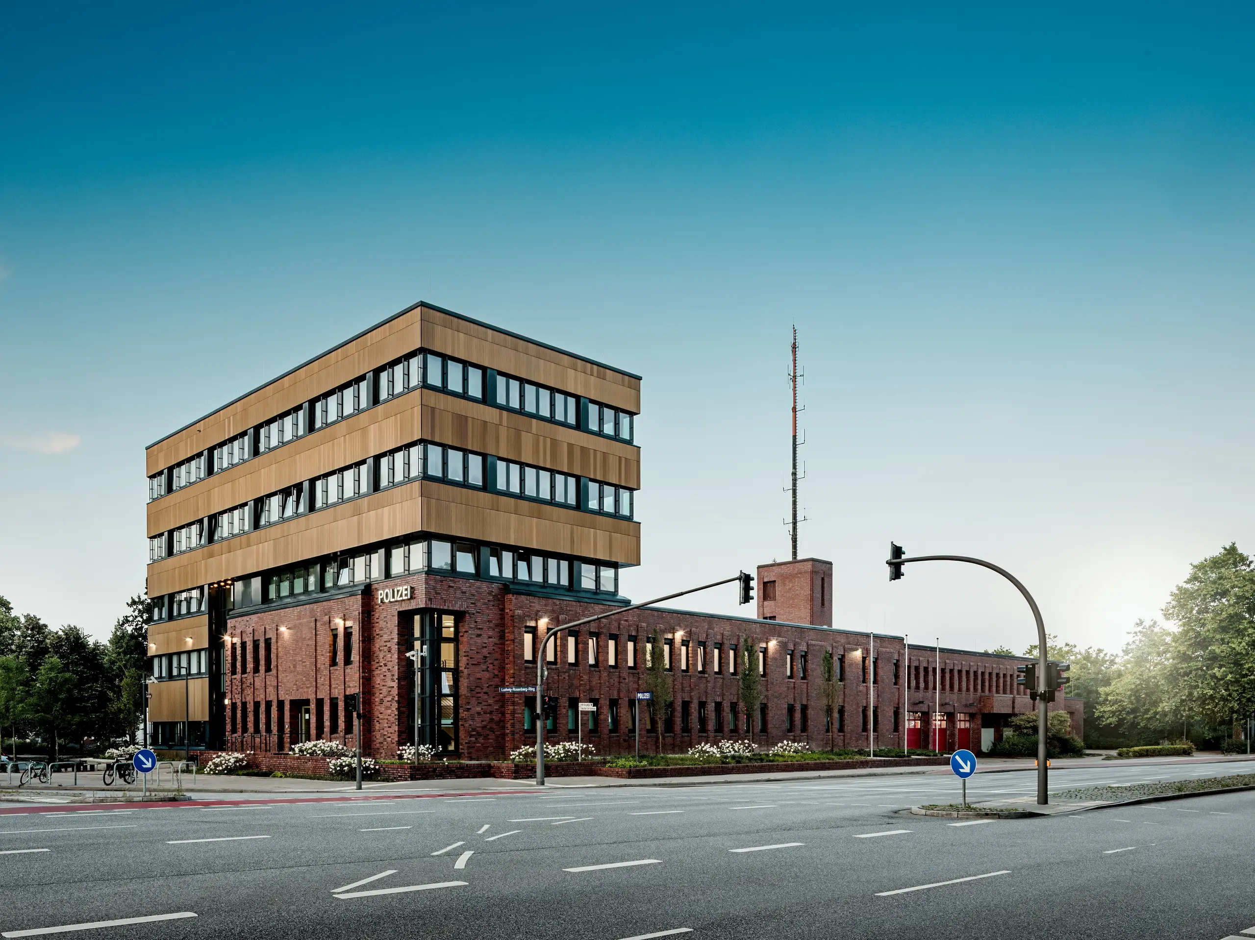 Weitläufige Straßenansicht des Polizeikommissariats 43 in Hamburg: Der präzise gegliederte Neubau erhebt sich über dem Backsteinbestand und fasziniert mit einer PREFA Aluminiumfassade aus Strangpressprofilen „Zacke“ in Mittelbronze. Die horizontale Schichtung und fein strukturierte Oberfläche erzeugen je nach Lichteinfall lebendige Reflexionen und machen das Gebäude zum architektonischen Eyecatcher. PREFA Aluminium überzeugt durch hohe Korrosionsbeständigkeit, Langlebigkeit und minimalen Wartungsaufwand – eine nachhaltige Lösung für moderne, funktionale und designstarke Fassaden.