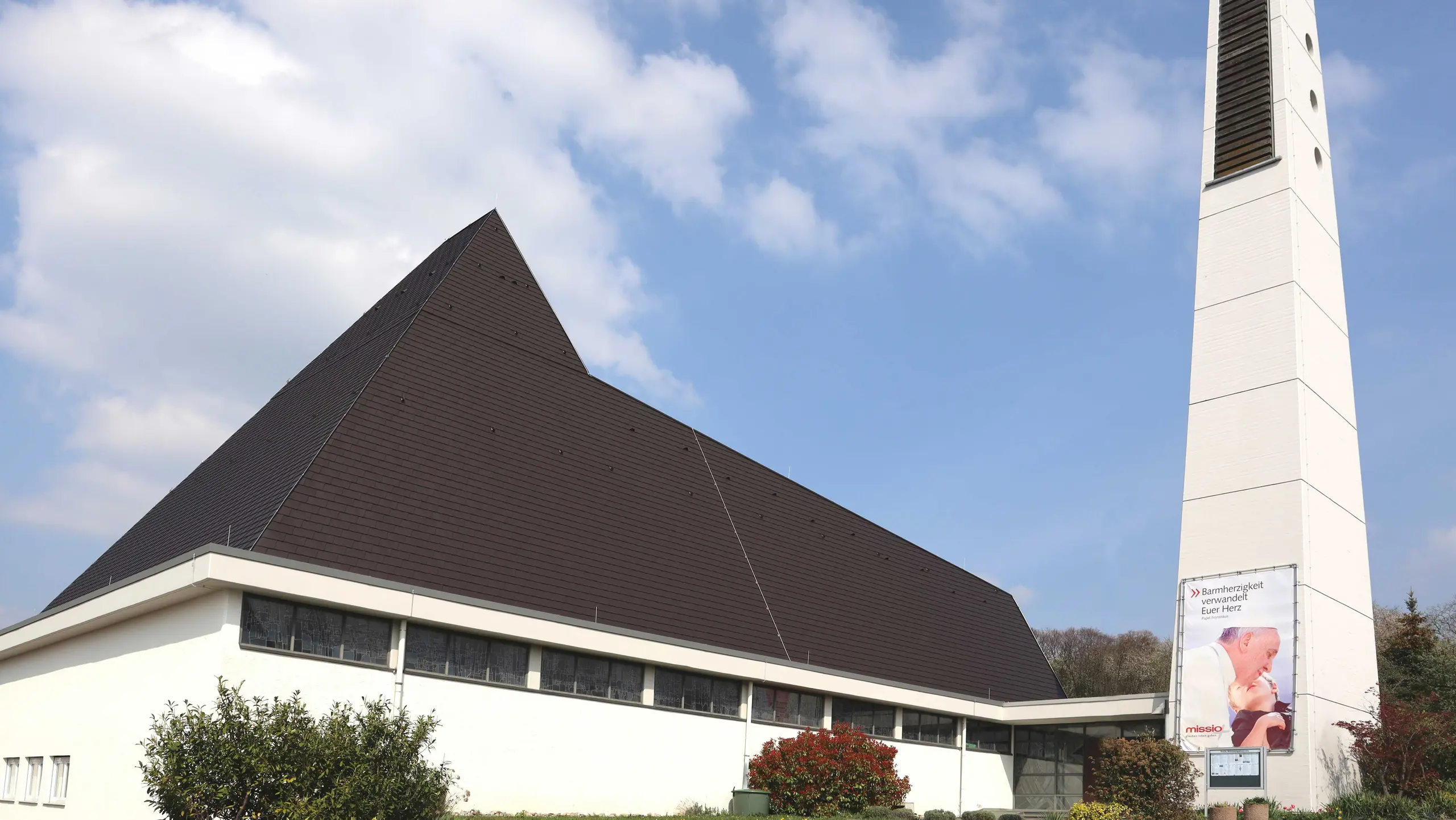 Kirchengebäude mit markantem, steil aufragendem Pyramidendach in dunklem Anthrazit und heller Putzfassade. Rechts steht ein hoher, heller Glockenturm mit vertikalen Öffnungen und einem Banner. Vor der Kirche befinden sich Sträucher und Blumenbeete, im Hintergrund ein blauer Himmel mit leichten Wolken.