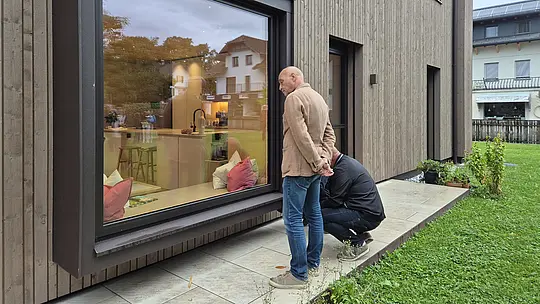 Zwei Teilnehmende der PREFA Objekttour begutachten aufmerksam die Ausführung eines modernen Einfamilienhauses mit vertikaler Holzfassade und großzügigem Panoramafenster. Der markante dunkle Fensterrahmen bildet einen starken architektonischen Eyecatcher. Hochwertige PREFA Fensterbänke und Aluminiumverblechungen setzen präzise Akzente und überzeugen durch ihre langlebige, wetterbeständige Ausführung.