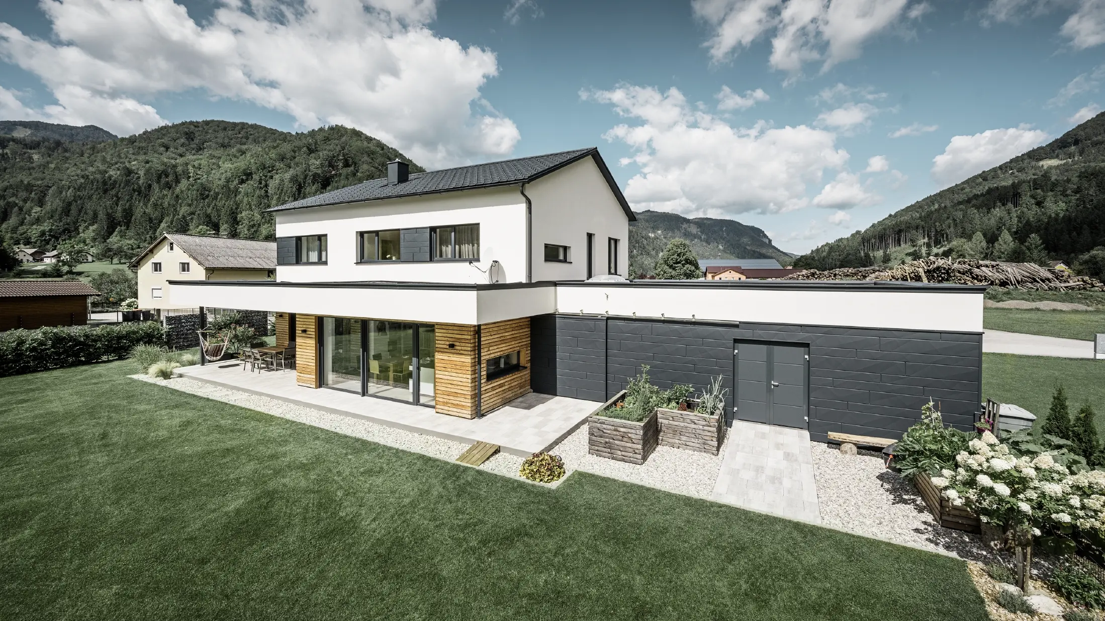 Das Bild zeigt ein modernes Einfamilienhaus in einer idyllischen Landschaft mit einer klaren Sicht auf bewaldete Hügel im Hintergrund. Das Haus zeichnet sich durch eine Kombination aus weißen Putzwänden und einer markanten Fassadenverkleidung mit PREFA Siding.X in P.10 Anthrazit aus, die für einen modernen und langlebigen Look sorgt. Die dunkle Färbung der Fassadenelemente bildet einen ansprechenden Kontrast zum hellen Außendesign und zur natürlichen Umgebung. Die Anordnung der Siding.X Fassadenpaneele ist präzise und verleiht der Immobilie einen strukturierten und hochwertigen Charakter, der typisch für PREFA Produkte ist. Das Dach des Gebäudes wurde mit der bewährten PREFA Dachplatte eingedeckt. Die Dachentwässerung erfolgt durch das Ablaufrohr und die Hängerinne von PREFA.