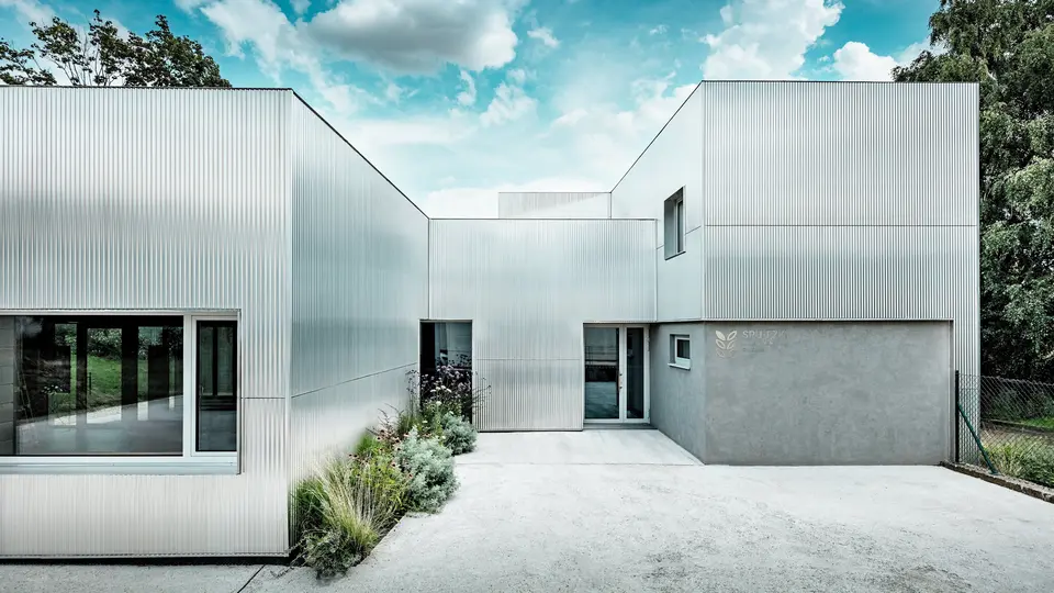 Das Envirocenter Nitra beeindruckt mit seiner silbern schimmernden PREFA Aluminiumfassade aus Strangpressprofil Welle in Naturblank. Die vertikalen Wellenstrukturen verbinden die unterschiedlich hohen, kubischen Baukörper zu einem harmonischen Ensemble. Großzügige Fenster, eine naturnahe Bepflanzung und der strahlend blaue Wolkenhimmel setzen spannende Akzente. Langlebige, witterungsbeständige Aluminiumprofile vereinen moderne Ästhetik und Funktionalität.