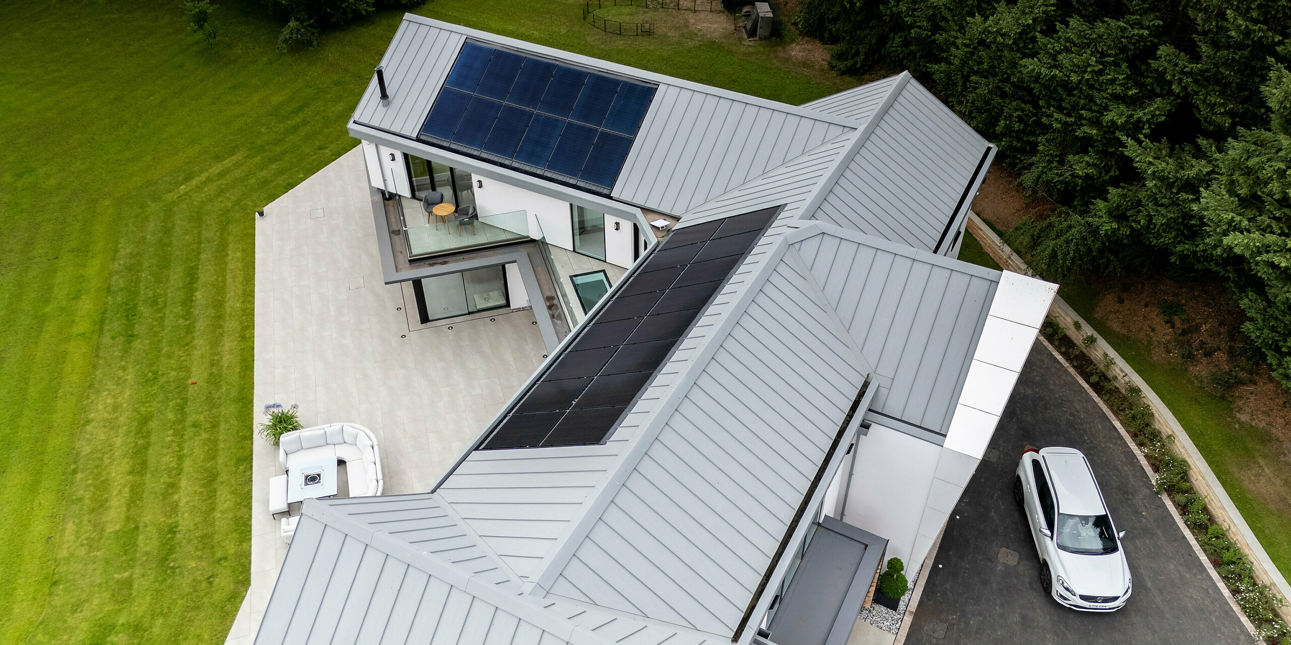 Die moderne Villa in Draycott in the Clay wird durch das elegante PREFALZ-Dach in der Farbe Patinagrau und die Photovoltaikanlage auf der dem Garten zugewandten Seite aufgewertet. Die klare Stehfalzstruktur verleiht ihr ein zeitloses, futuristisches Design, das perfekt zur grünen Umgebung passt. Das hochwertige Blechdach ist nicht nur ein ästhetischer Hingucker, sondern unterstützt auch die Energieeffizienz der Immobilie. Die Kombination aus hochwertigem Metall und Solartechnologie macht dieses Zuhause zu einem Vorreiter in puncto nachhaltiges und stilvolles Bauen. Vor dem Gebäude parkt ein weißes Auto, das auf Besucher schließen lässt.