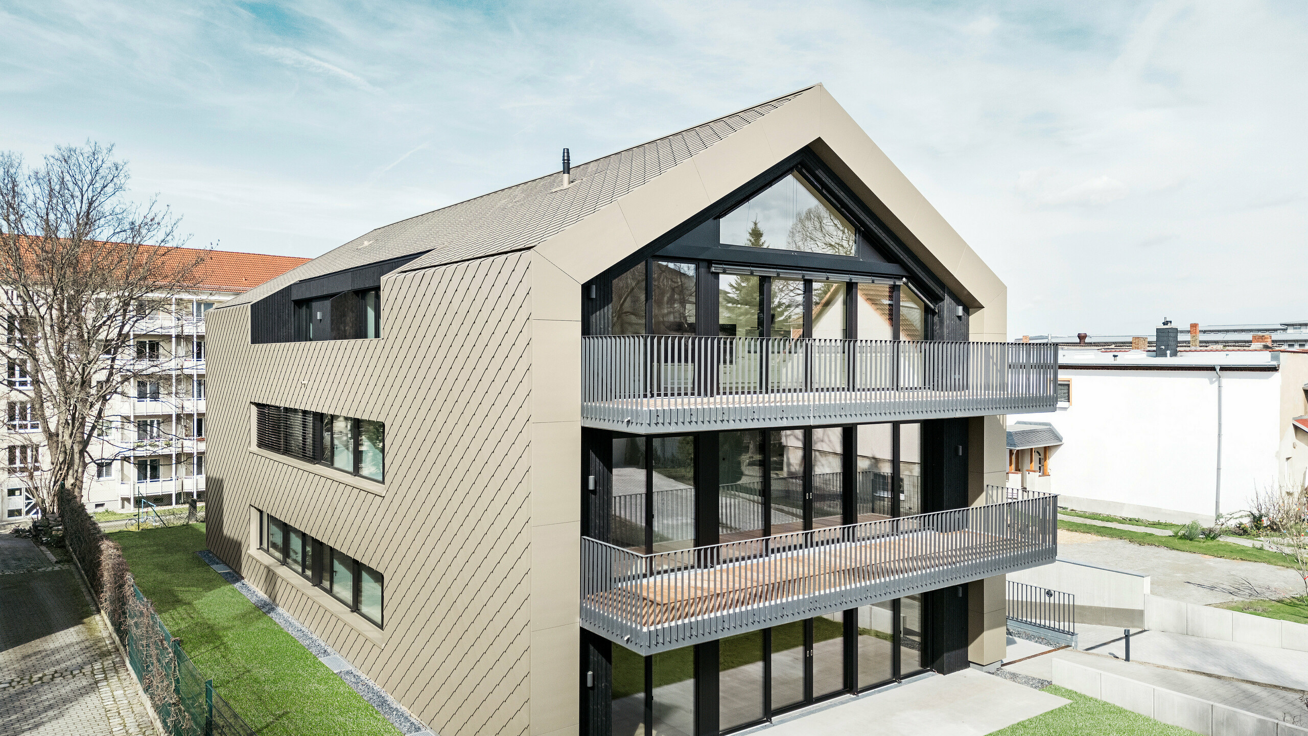 Gartenansicht eines modernen Mehrfamilienhauses in der Chemnitzer Straße in Dresden. Der gepflegte Garten liegt vor einer eindrucksvollen Architektur. Die Fassade und das Dach des Gebäudes sind mit der langlebigen PREFA Aluminiumraute 29 × 29 in Bronze verkleidet, die dem Bauwerk eine elegante und zeitgemäße Optik verleiht. Das Bild zeigt die großzügigen Balkone, die großen Fensterfronten und die harmonische Einbettung des Gebäudes in die Umgebung.