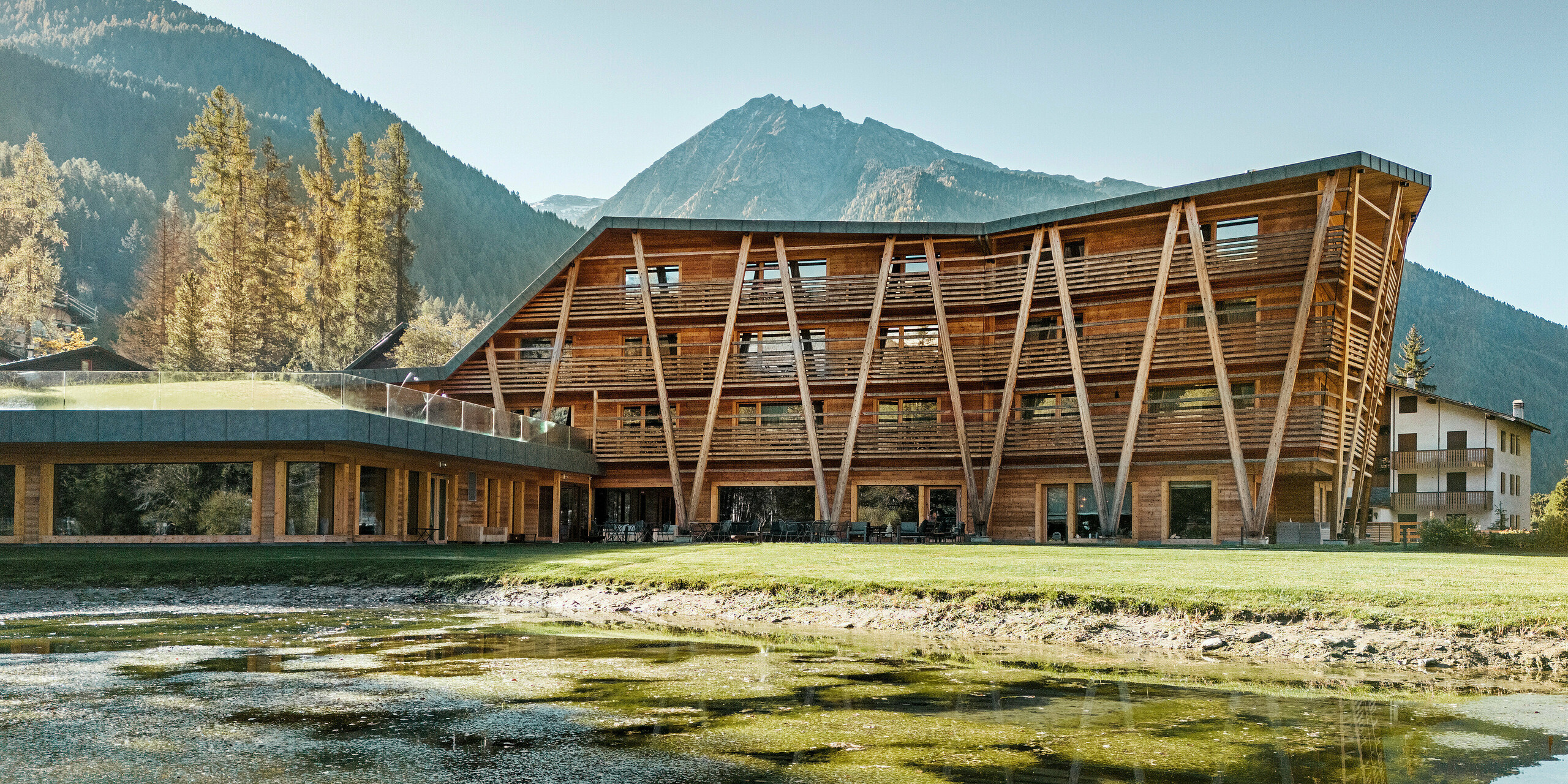 Das Bild zeigt die beeindruckende Pracht des 'Au Charmant Petit Lac', eines Luxus-Öko-Hotels in Champoluc, Italien, vor dem Hintergrund majestätischer Berge. Das Hotel zeichnet sich durch eine ausdrucksstarke Holzarchitektur aus, die sich nahtlos in die natürliche Umgebung einfügt. Der kleine See im Vordergrund spiegelt die ruhige und entspannende Atmosphäre wider, die das Hotel umgibt. Die umweltbewusste Bauweise wird durch die Nutzung von PREFALZ in P.10 Steingrau für das Dach unterstrichen, was die nachhaltige Philosophie des 5-Sterne-Hauses betont.