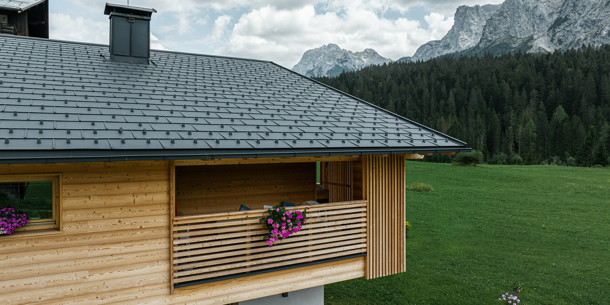 Die malerische Seitenansicht der Loggia von Casa Hoffe in Sappada, Italien, zeigt die harmonische Integration von PREFA Dachplatten R.16 in P.10 Schwarz mit einer naturbelassenen Holzfassade. Die traditionelle Bauweise der Loggia, kombiniert mit der modernen, langlebigen Dachlösung, fügt sich nahtlos in die atemberaubende Bergkulisse der Dolomiten ein und unterstreicht das naturnahe Wohnkonzept der Region.