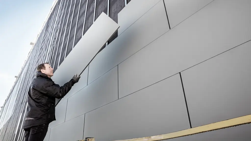 Fotos einer teilweise fertiggestellten Fassade, die mit PREFA Sidings in der Breite 500 verkleidet ist. Der Spengler verlegt ein weiteres Fassadenpaneel. Das Bild ist von unten aufgenommen. 