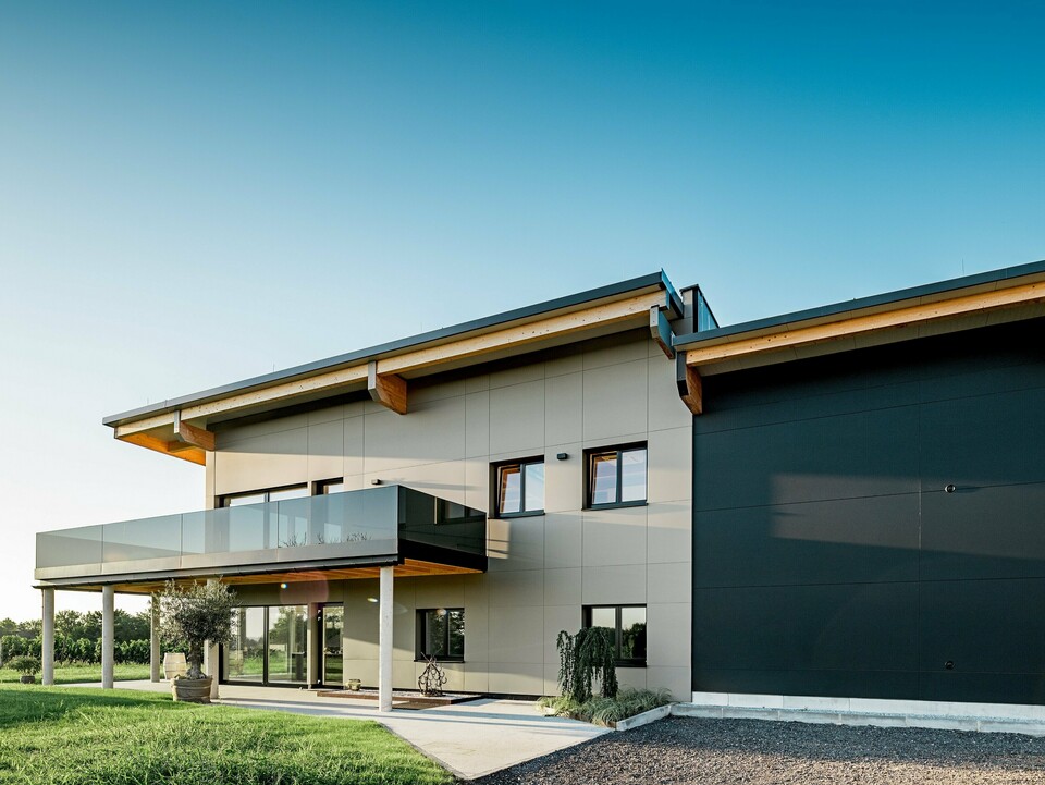 Seitenansicht des Weinguts Nigl in Perchtoldsdorf mit kontrastreicher PREFA Aluminiumfassade in den Farben P.10 Prefabronze und P.10 Schwarz. Links dominiert die helle Fassadenseite mit großzügigem Balkon samt Glasbrüstung und filigranen Stützen, während rechts die tiefschwarze Aluminiumfläche mit klaren Linien Ruhe und Stärke ausstrahlt. Sichtbare Holzdachträger unterstreichen den modernen Landhausstil. Die Kombination aus hochwertigen PREFA Verbundplatten, Glas, Holz und Beton schafft eine zeitlose, elegante Architektur mit maximaler Witterungsbeständigkeit und Langlebigkeit – perfekt für repräsentative Bauten im Weinbauumfeld.