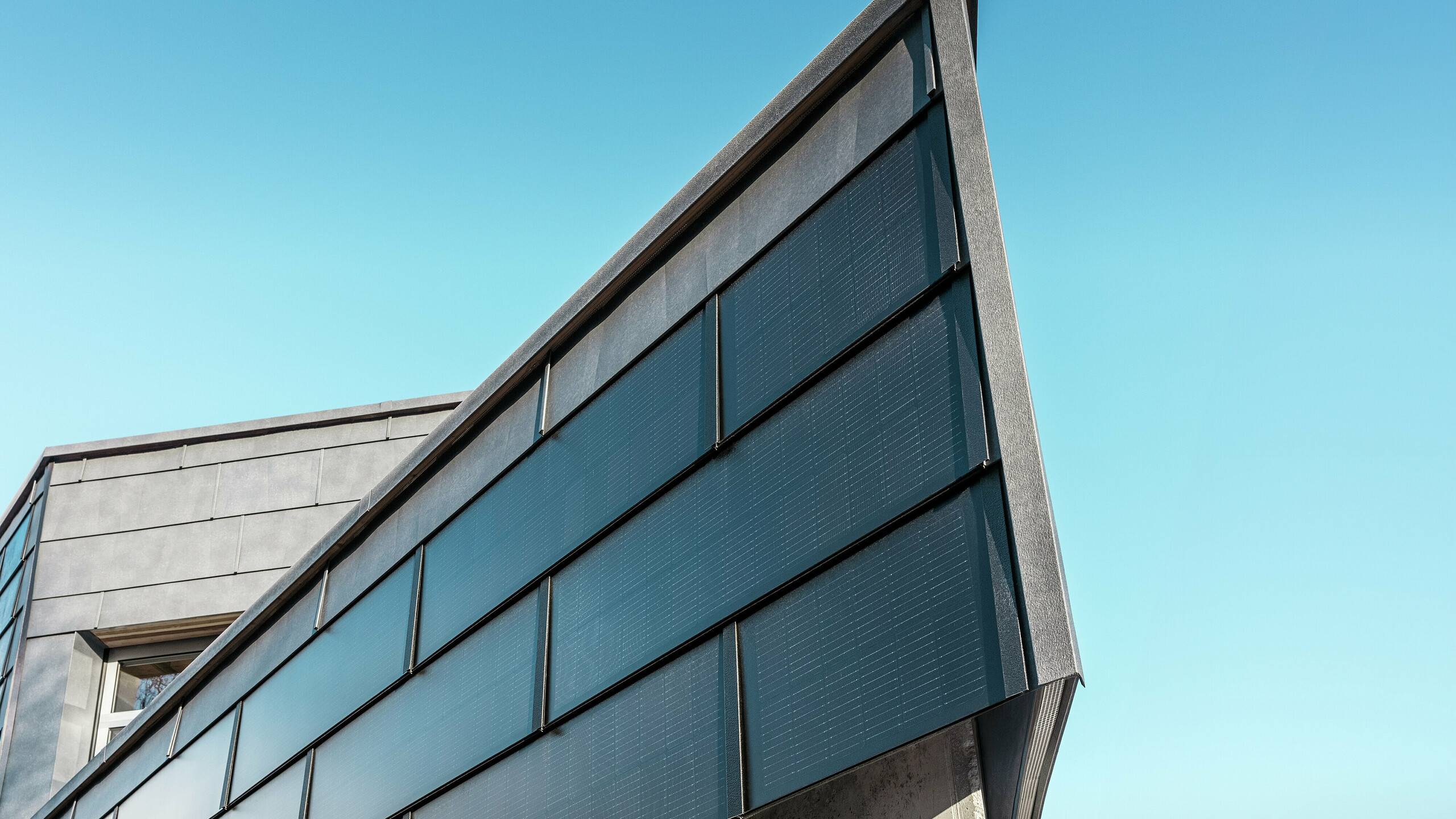 Moderne Firmenfassade der Spenglerei RÜMA in Vorarlberg mit PREFA Solardachplatten aus Aluminium und integrierten Photovoltaikmodulen, kombiniert mit PREFA Fassadenpaneelen FX.12 für nachhaltige Energiegewinnung und dynamisches Fassadendesign. Die Aufnahme zeigt die Gebäudeecke aus nächster Nähe. Im Hintergrund ist der strahlend blaue Himmel zu sehen. 