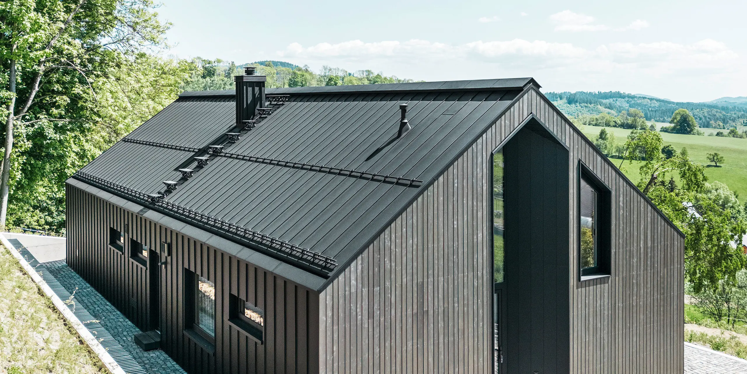 A dark solid-timber house clad in PREFALZ in P. 10 on the roof and the reverse of the building. In front of the house stands a small outbuilding with an aluminium cladding of PREFALZ in P.10 black. In the background, a bright blue sky and woodland.