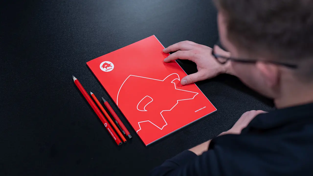 Blick auf einen Tisch mit PREFA-Schreibmaterialien, darunter ein rotes Notizbuch mit dem ikonischen PREFA-Logo und passende rote Stifte. Eine Person steht kurz davor, das Notizbuch zu öffnen, was auf die wertvollen Informationen und Ressourcen der PREFA Academy hinweist. Das Bild verweist auf den Infobereich der Academy, der Fachwissen und Schulungsmaterialien für Architekten und Fachleute bietet.
