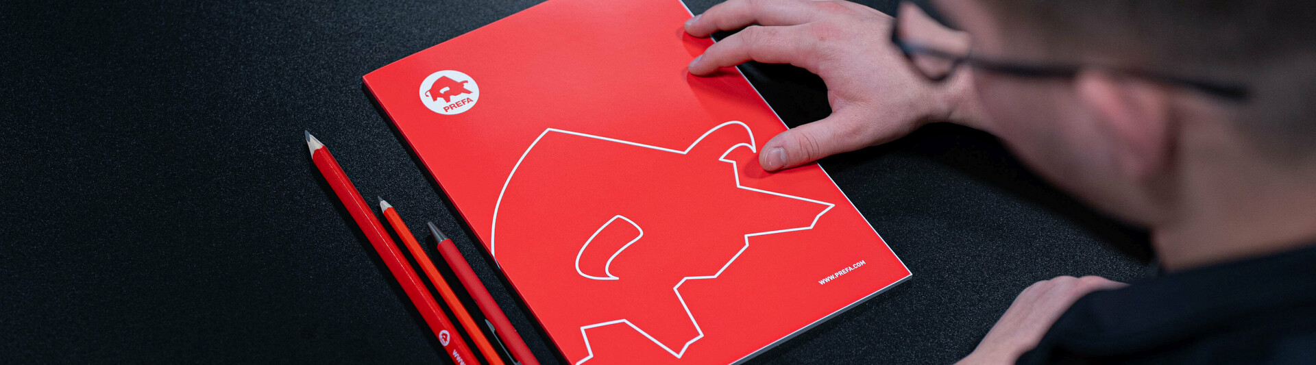 Blick auf einen Tisch mit PREFA-Schreibmaterialien, darunter ein rotes Notizbuch mit dem ikonischen PREFA-Logo und passende rote Stifte. Eine Person steht kurz davor, das Notizbuch zu öffnen, was auf die wertvollen Informationen und Ressourcen der PREFA Academy hinweist. Das Bild verweist auf den Infobereich der Academy, der Fachwissen und Schulungsmaterialien für Architekten und Fachleute bietet.