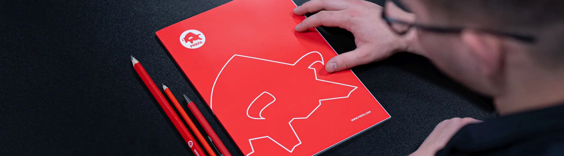 Blick auf einen Tisch mit PREFA-Schreibmaterialien, darunter ein rotes Notizbuch mit dem ikonischen PREFA-Logo und passende rote Stifte. Eine Person steht kurz davor, das Notizbuch zu öffnen, was auf die wertvollen Informationen und Ressourcen der PREFA Academy hinweist. Das Bild verweist auf den Infobereich der Academy, der Fachwissen und Schulungsmaterialien für Architekten und Fachleute bietet.