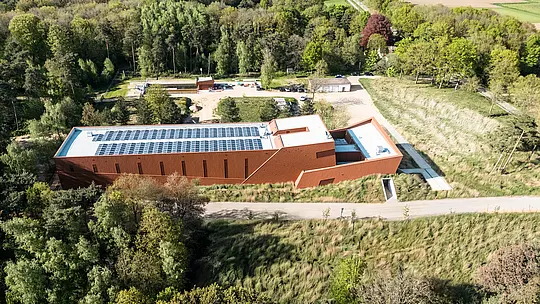 Luftaufnahme des Trinkwasserpumpwerks WML in Lottum als markanter Infrastrukturbau, harmonisch eingebettet in eine grüne Wald- und Wiesenlandschaft. Der langgestreckte Baukörper mit prägnanter PREFA Siding.X Aluminiumfassade in warmer Rostoptik wirkt wie eine skulpturale Landmarke im Gelände. Eyecatcher sind die klar strukturierte Gebäudeform, die integrierten Photovoltaikmodule auf dem Dach sowie die fein facettierte Fassadenoberfläche. Die langlebigen, leichten und recycelbaren PREFA Aluminium-Paneele überzeugen durch hohe Witterungsbeständigkeit, nachhaltige Effizienz und eine wartungsarme Gebäudehülle.