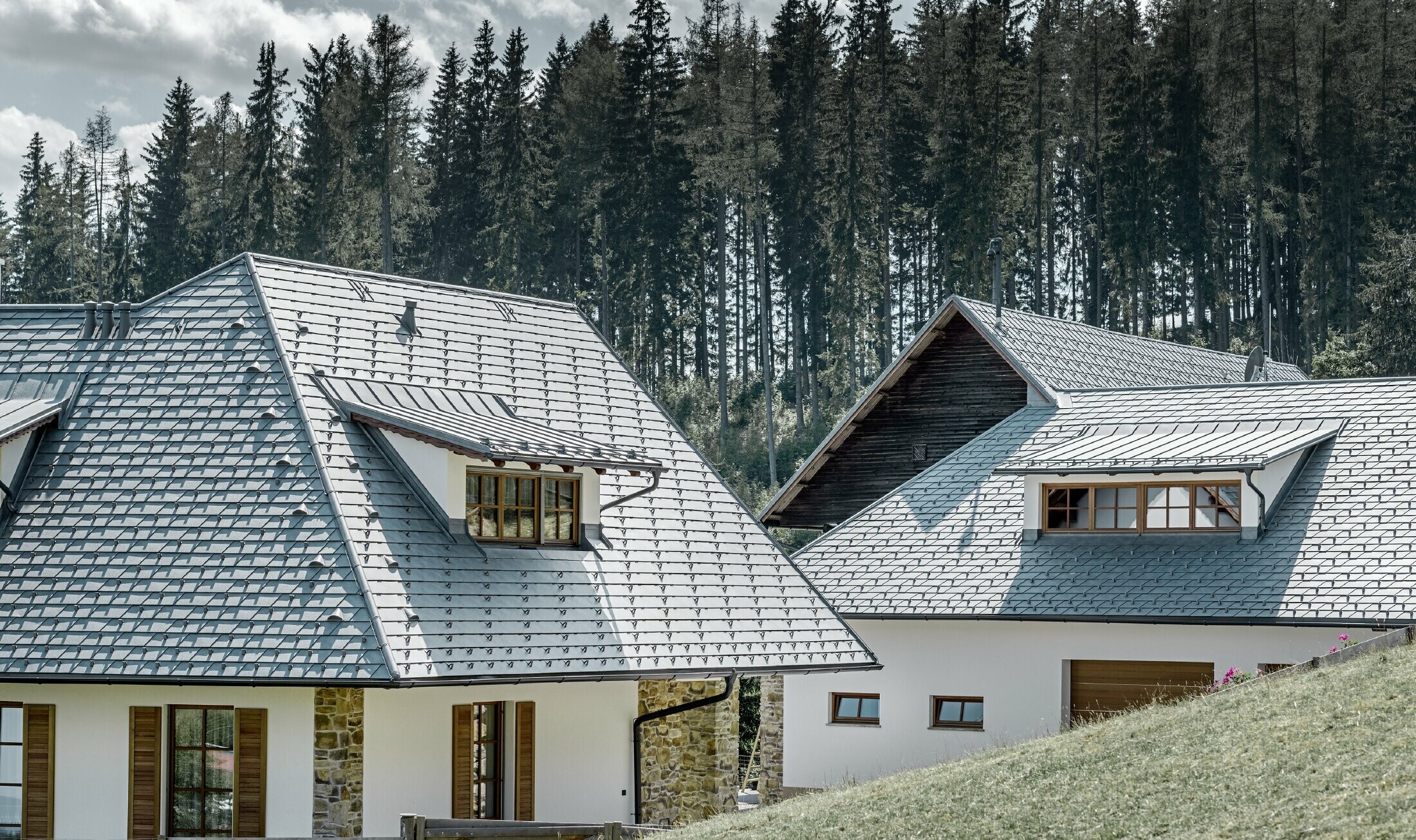Rückansicht des Einfamilienhausees in Hanglage; Das Dach wurde mit der Alu-Dachschindel in Steingrau eingedeckt, die Schleppgauben mit Stehalz, ebenfalls in Steingrau. In Hintergrund erkennt man einen Wald.