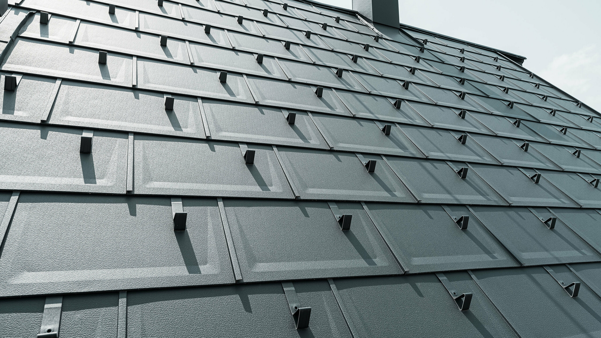 Detailaufnahme der PREFA Dachplatte R.16 in der Farbe P.10 Anthrazit. Am Dach sind PREFA Schneestopper und ein Rauchfang zu sehen.