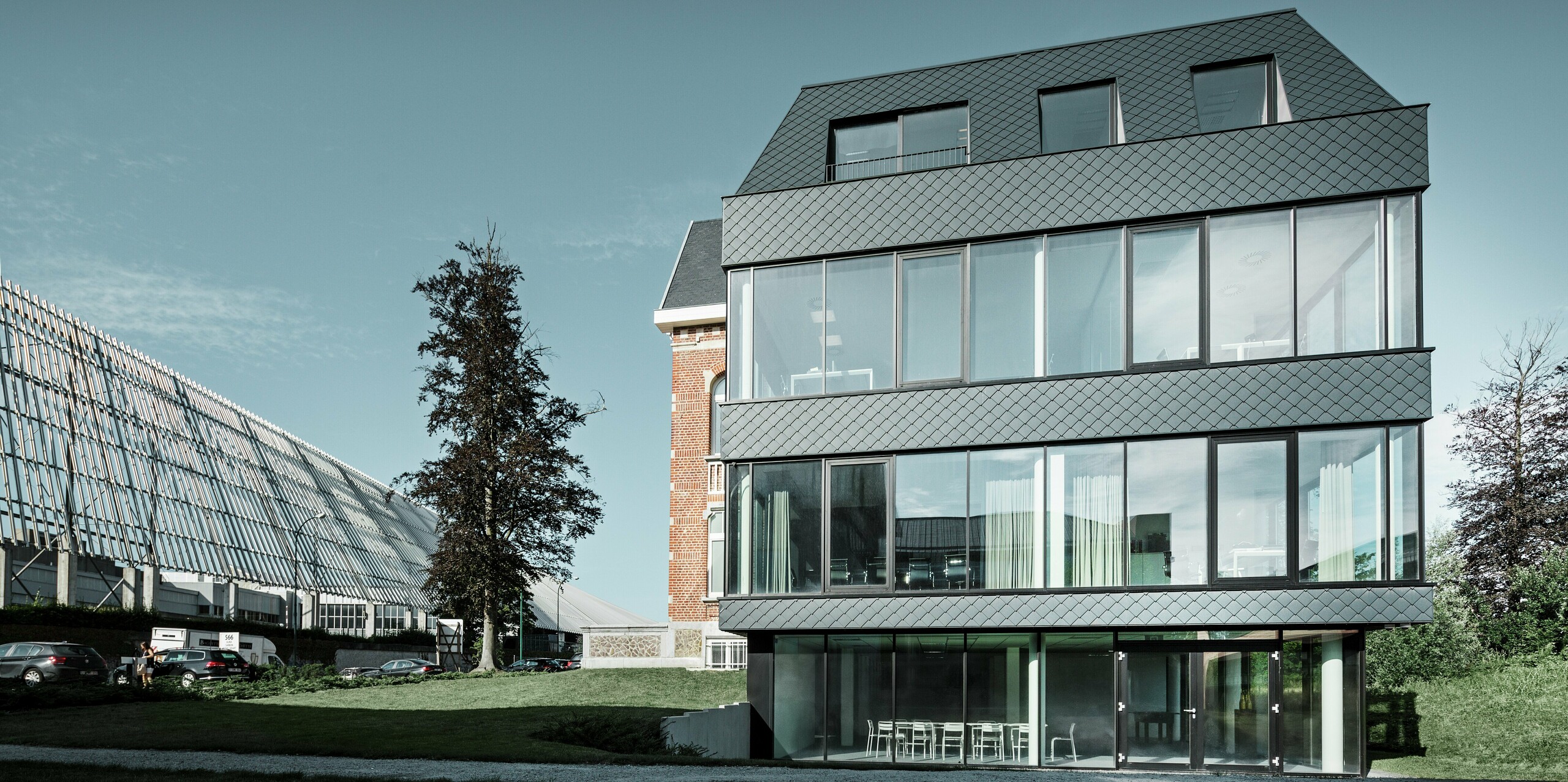 Frontalansicht des modernen Zubaus am Kasteeltje Heizel: Großzügige Glasfronten öffnen den Blick ins Innere, während die anthrazitfarbenen PREFA Wandrauten dem Gebäude eine elegante, strukturierte Oberfläche verleihen. Im Zusammenspiel mit der historischen Backsteinvilla entsteht ein architektonisches Highlight, das Moderne und Tradition in nachhaltiger Leichtigkeit vereint.