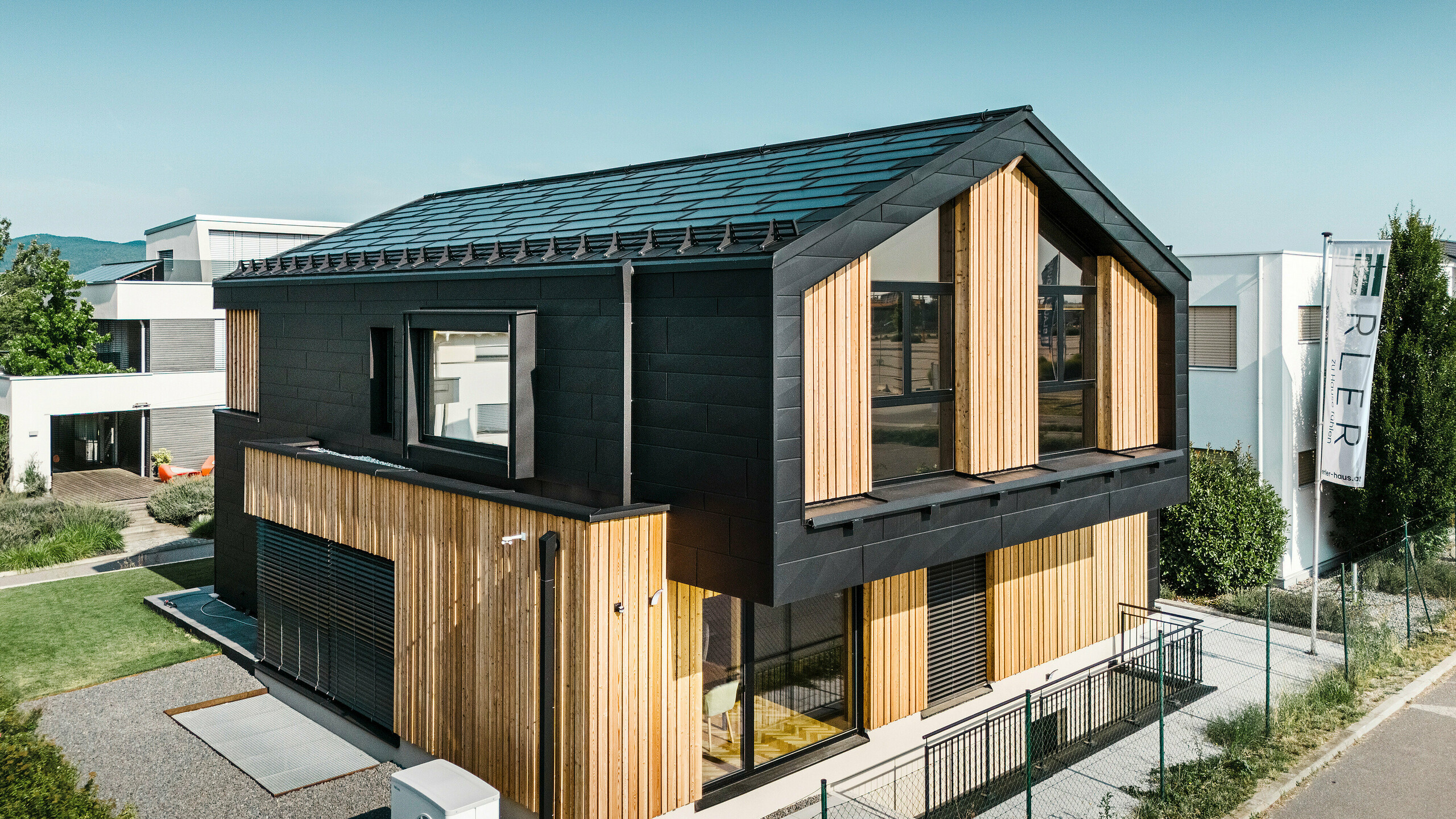 Vorderseite des Musterhauses Autark im Fertighauszentrum Blaue Lagune, Vösendorf (AT). Das Dach ist mit schwarzen PREFA Solardachplatten und Dachplatten R.16 in hochwertiger P.10-Beschichtung eingedeckt. Die Fassade besteht aus einer Kombination von vertikalen Holzelementen und PREFA Siding.X in P.10 Schwarz, was dem Gebäude einen modernen und nachhaltigen Charakter verleiht. Am rechten Bildrand weht die Fahne der Erler Bau GmbH.