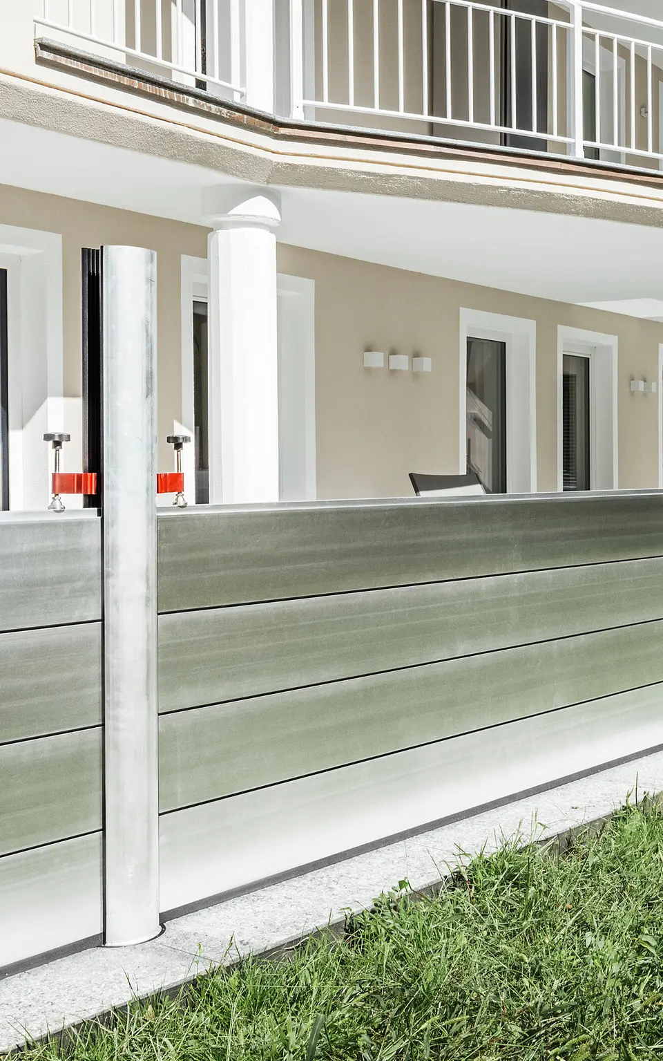 Panoramaansicht eines exklusiven Wohnhauses in Tirol mit installiertem PREFA Hochwasserschutzsystem entlang der Terrasse. Die Dammbalken aus Aluminium verlaufen über die gesamte Gebäudelänge und schützen mehrere Zugänge wie Terrassentüren und Fenster vor möglichem Hochwasser aus dem angrenzenden Gelände. Die moderne Architektur des Hauses mit Rundbalkon und weißen Stützen bildet einen stilvollen Kontrast zum funktionalen Hochwasserschutz aus Aluminium.