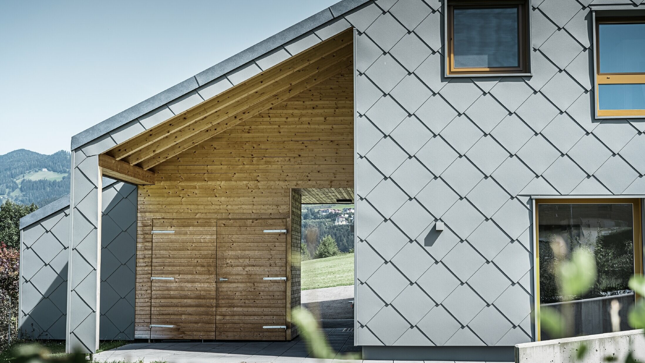 Der überdachte Eingangsbereich ist mit waagrechter Holzverkleidung gemacht, der Rest der Fassade ist mit der großen PREFA Wandraute aus Aluminium in Hellgrau verkleidet