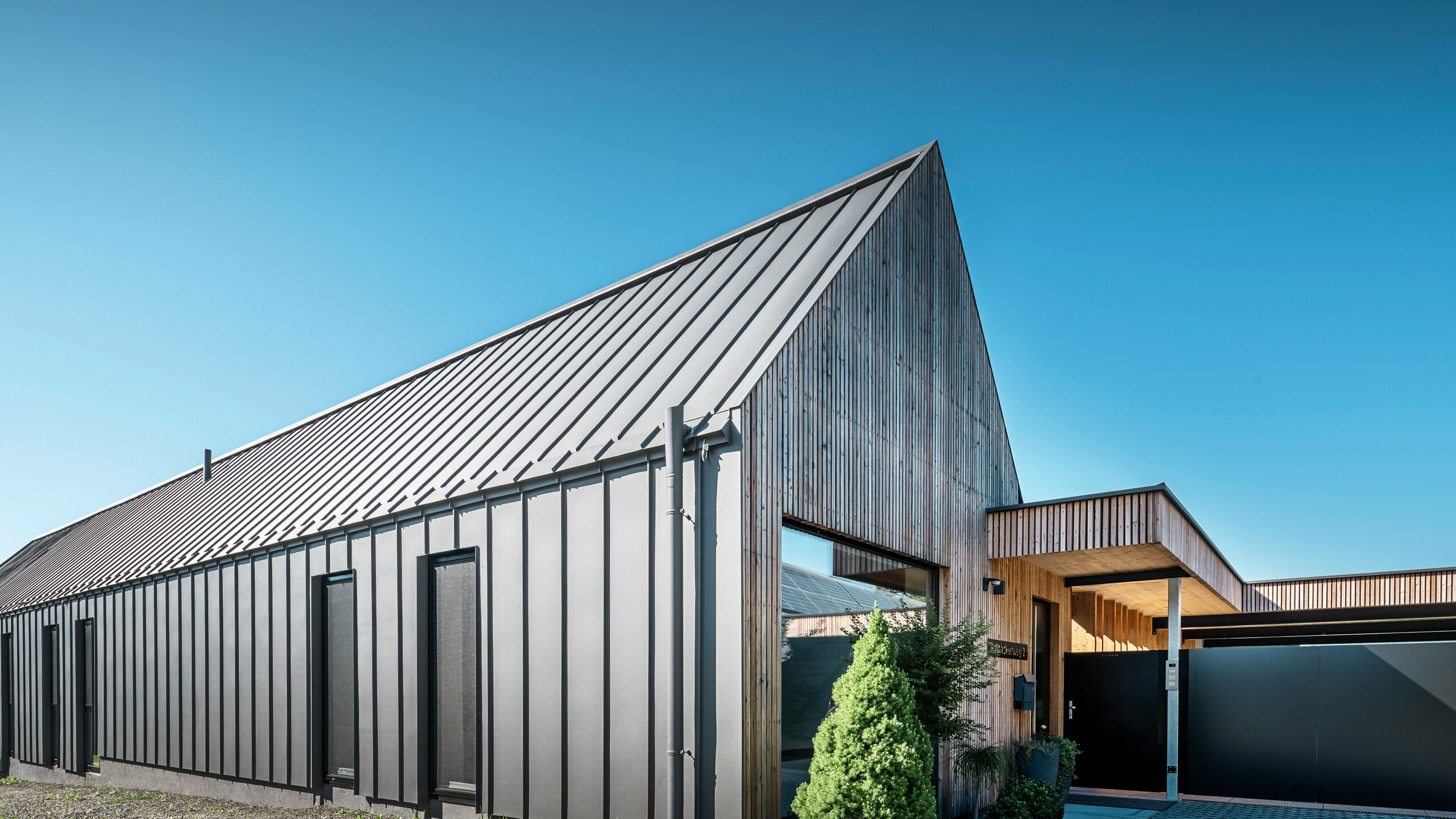 Eingangsbereich eines Einfamilienhauses mit angrenzender Garage in Pogier, Österreich, inmitten einer malerischen Landschaft. Dach und Fassade beider Gebäude sind mit langlebigem PREFALZ in dunkelgrau P.10 eingedeckt, was ein modernes Erscheinungsbild ergibt. Die Garage hat eine harmonisch integrierte Holzverkleidung, die sich nahtlos in die umgebende Landschaft einfügt. Der gepflasterte Hof und die klaren Linien des Entwurfs runden die Ästhetik des Grundstücks ab, während die sanften Hügel im Hintergrund das Bild einer ruhigen grünen Oase im grünen Herzen Österreichs vervollständigen.