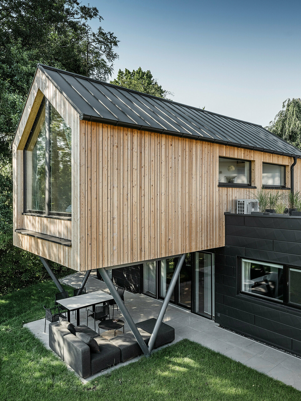 Das 'Woodstock Chalet 5' beeindruckt mit einer eindrucksvollen Kombination aus natürlichen Holzoberflächen und den modernen PREFA Siding.X Fassadenelementen in Anthrazit. Die überhängende Holzstruktur des Obergeschosses ragt markant über die dunklen Fassadenelemente, die das Erdgeschoss verkleiden, und erzeugt so ein Spiel aus Licht und Schatten. Die klaren Linien und die robuste Konstruktion der Siding.X Elemente ergänzen das innovative Design des Gebäudes und bieten einen spannenden visuellen Kontrast zur umgebenden Vegetation und dem traditionellen Gebäude im Hintergrund. Am Dach des Chalets wurde das PREFALZ Dachsystem in Form einer gleichmäßigen Stehfalzdeckung verlegt. Am rechten Rand des Erdgeschosses ist das PREFA Dachentwässerungssystem zu erkennen, das analog zur Aluminiumfassade in Anthrazit gehalten ist. Für die Dachentwässerung wurden der Wasserfangkasten gefolgt vom Ablaufrohr montiert.