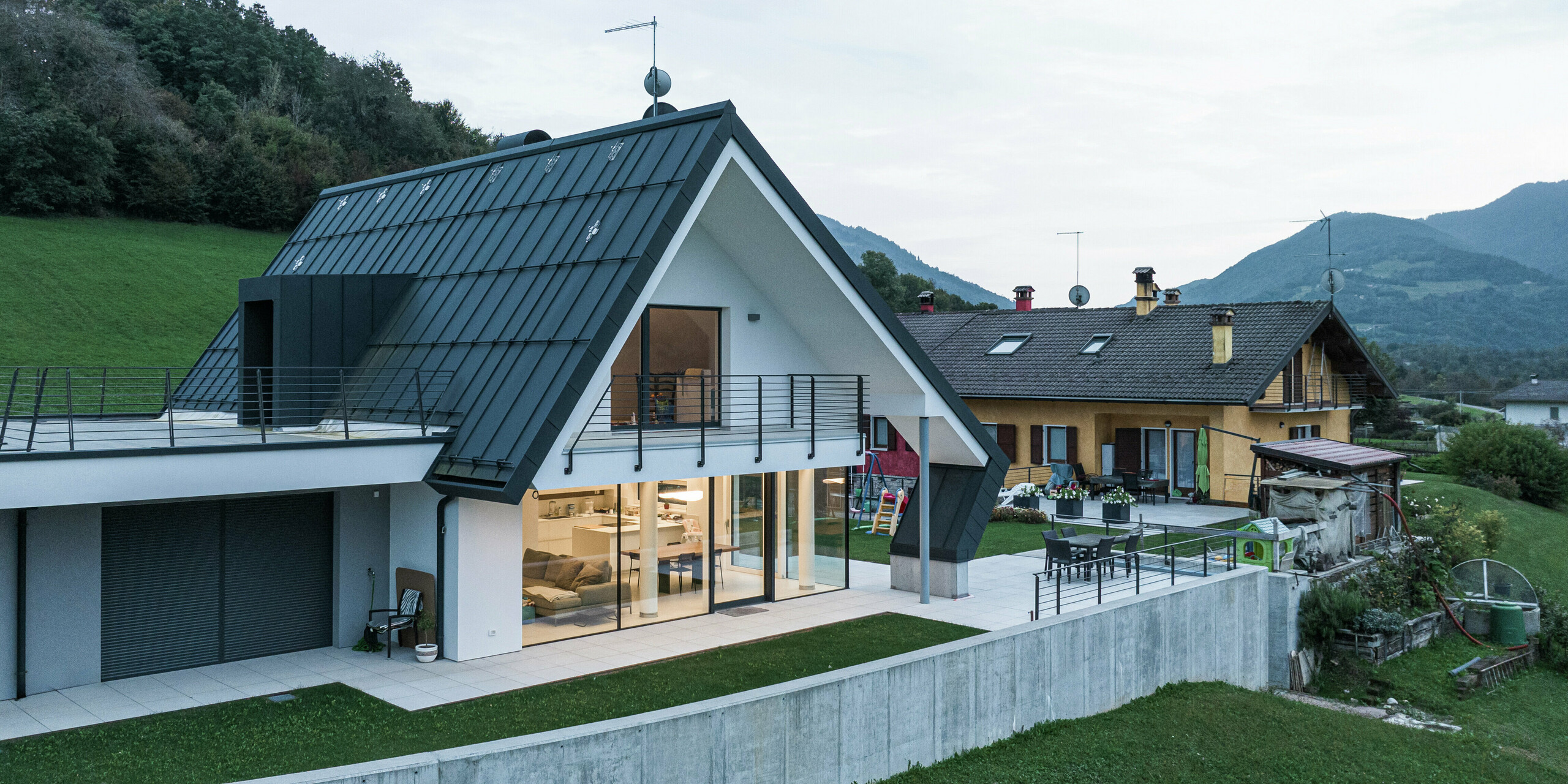 Modernes Einfamilienhaus mit Blick auf Garten und Terrasse. Das Aluminiumdach mit markanten Dachüberständen schützt die Fassade und schafft einen harmonischen Übergang zur großzügigen Terrasse. Große Fensterflächen sorgen für lichtdurchflutete Räume, der umlaufende Balkon unterstreicht den offenen Wohncharakter. Die Kombination aus langlebigem PREFA Aluminium, moderner Architektur und hochwertiger Verarbeitung macht dieses Haus besonders langlebig und stilvoll.