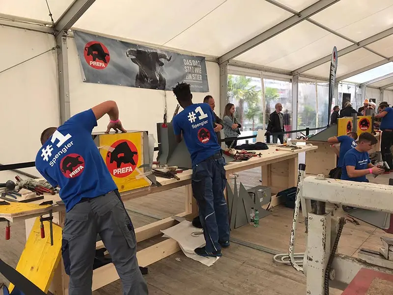 Spengler-Lehrlinge verarbeiten das Aluminium-Falzmaterial Prefalz. Besucher besichtigen die erstellten Kamineinfassungen.