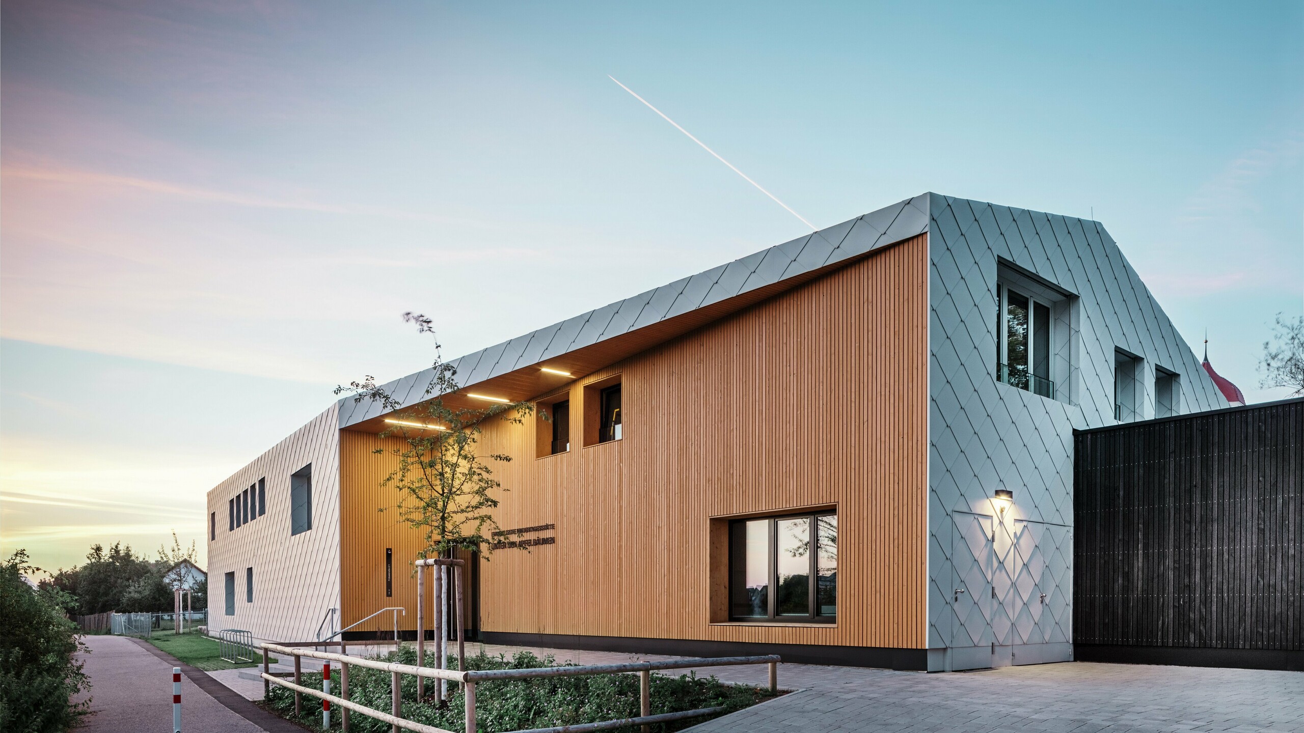 Aufnahme bei Dämmerung einer modernen Kita in Ulm. Das Dach wurde ohne Dachvorsprung ausgeführt. Der Großteil der Fassade wurde mit großflächigen Aluminium Rauten von PREFA in Weiss Verkleidet, dazu wurde der Eingangsbereich großflächig mit vertikalen Holzpaneelen verkleidet.