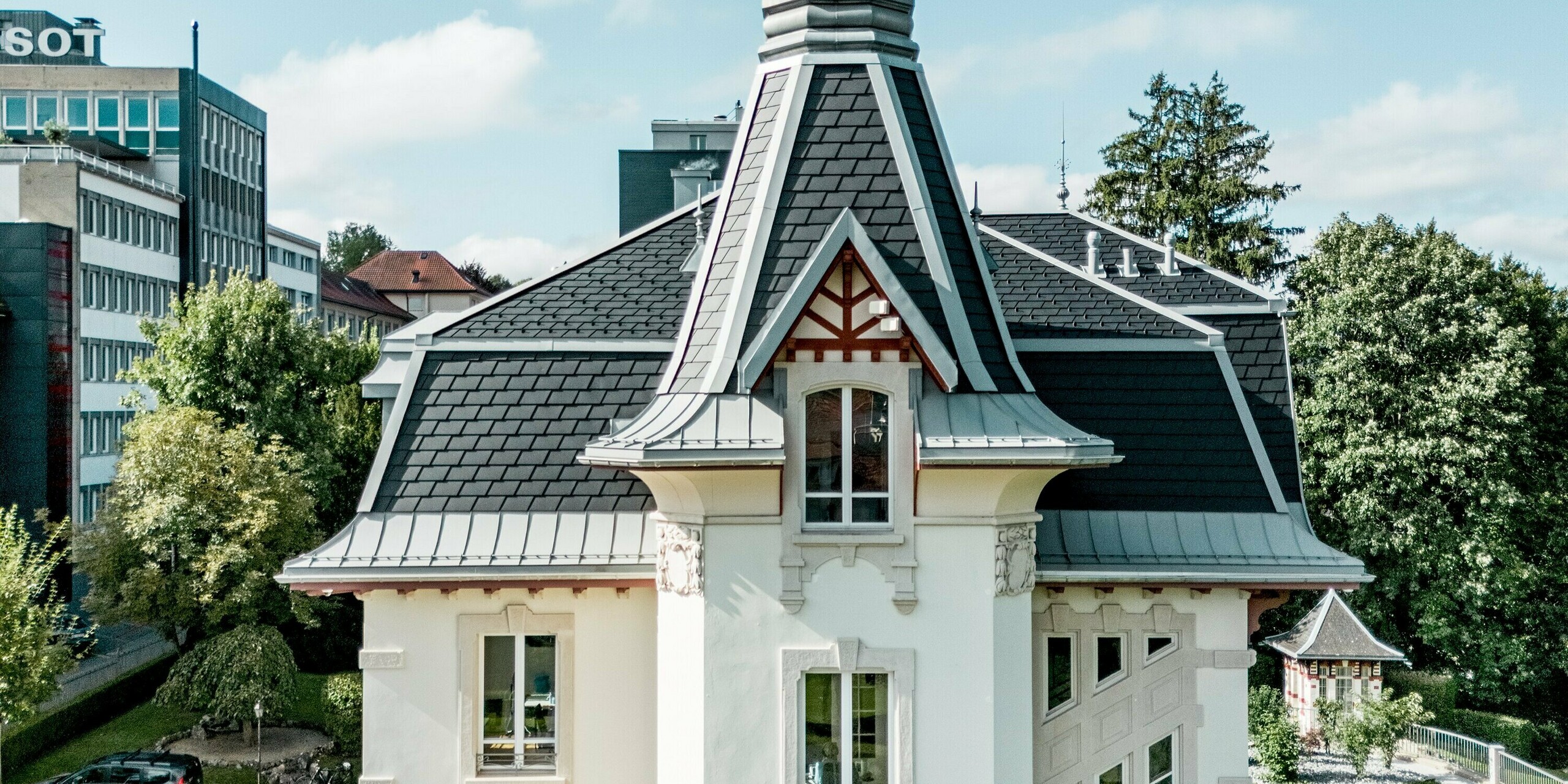 Markante Seitenansicht der Manufacture Horlogère du Locle in der Schweiz mit ikonischem Turm als Eyecatcher, der elegant in den Himmel ragt. Die komplexe Dachlandschaft fasziniert durch steil geneigte Flächen und präzise gegliederte Geometrie aus PREFA Dachschindeln in P.10 Schwarz. PREFALZ in Patinagrau setzt geschwungene Übergänge und feine Details perfekt um. Das langlebige, leichte Aluminiumdach überzeugt durch hohe Widerstandsfähigkeit, maximale Gestaltungsfreiheit und schont gleichzeitig die historische Bausubstanz nachhaltig.