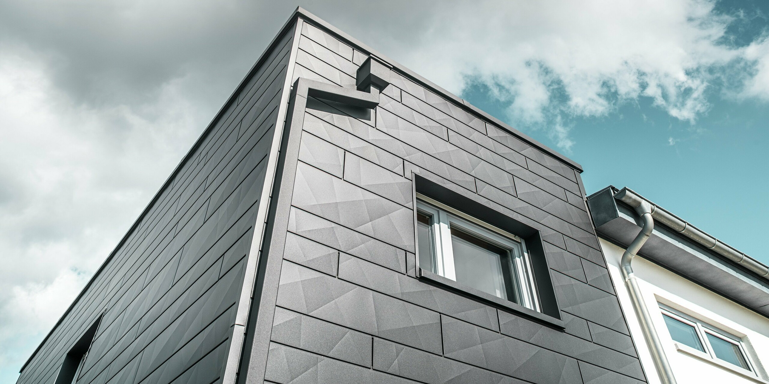 Die markante Fassade des sanierten Einfamilienhauses in Teuschnitz zeigt die PREFA Siding.X Aluminiumpaneele in P.10 Schwarz, die eine moderne und geometrische Struktur schaffen. Die Fassadenelemente verlaufen in klaren Linien und betonen den zeitlosen Charakter des Gebäudes. Ein Fenster wird von schwarzen Aluminium Verbundplatten umrahmt. Die integrierte PREFA Dachentwässerung mit dem Quadratrohr und dem Wasserfangkasten fügt sich harmonisch in das Gebäude ein und sorgt für eine stilvolle und funktionale Lösung.