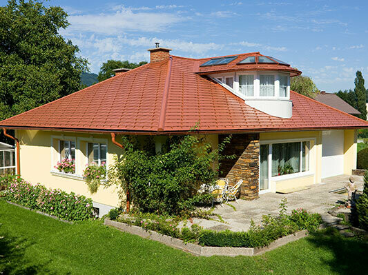Rodinný dům s valbovou střechu a vikýřem pokryté PREFA hliníkovými šindely v cihlově červené.