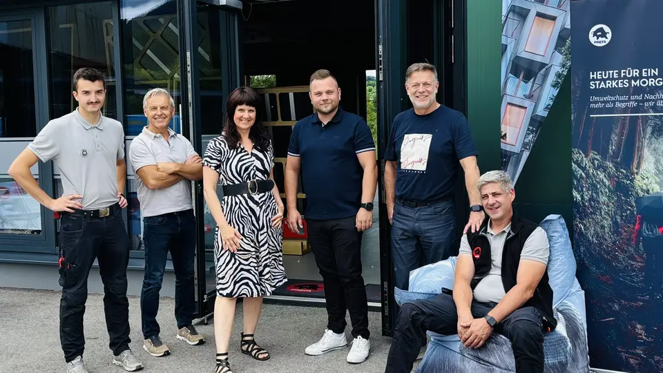 Gruppenfoto vor dem PREFA Showroom in St. Veit an der Glan: Mehrere PREFA Mitarbeiter stehen und sitzen gemeinsam vor dem Eingang. Neben der geöffneten Tür ist ein PREFA Banner mit Architekturprojekten und Aluminium-Dach- und Fassadenlösungen sichtbar. Die moderne Gebäudefassade und der Eingangsbereich unterstreichen den professionellen Rahmen. Die Szene zeigt den persönlichen Austausch, das Netzwerk sowie die gelungene Veranstaltung rund um PREFA Aluminiumprodukte für Dach und Fassade.