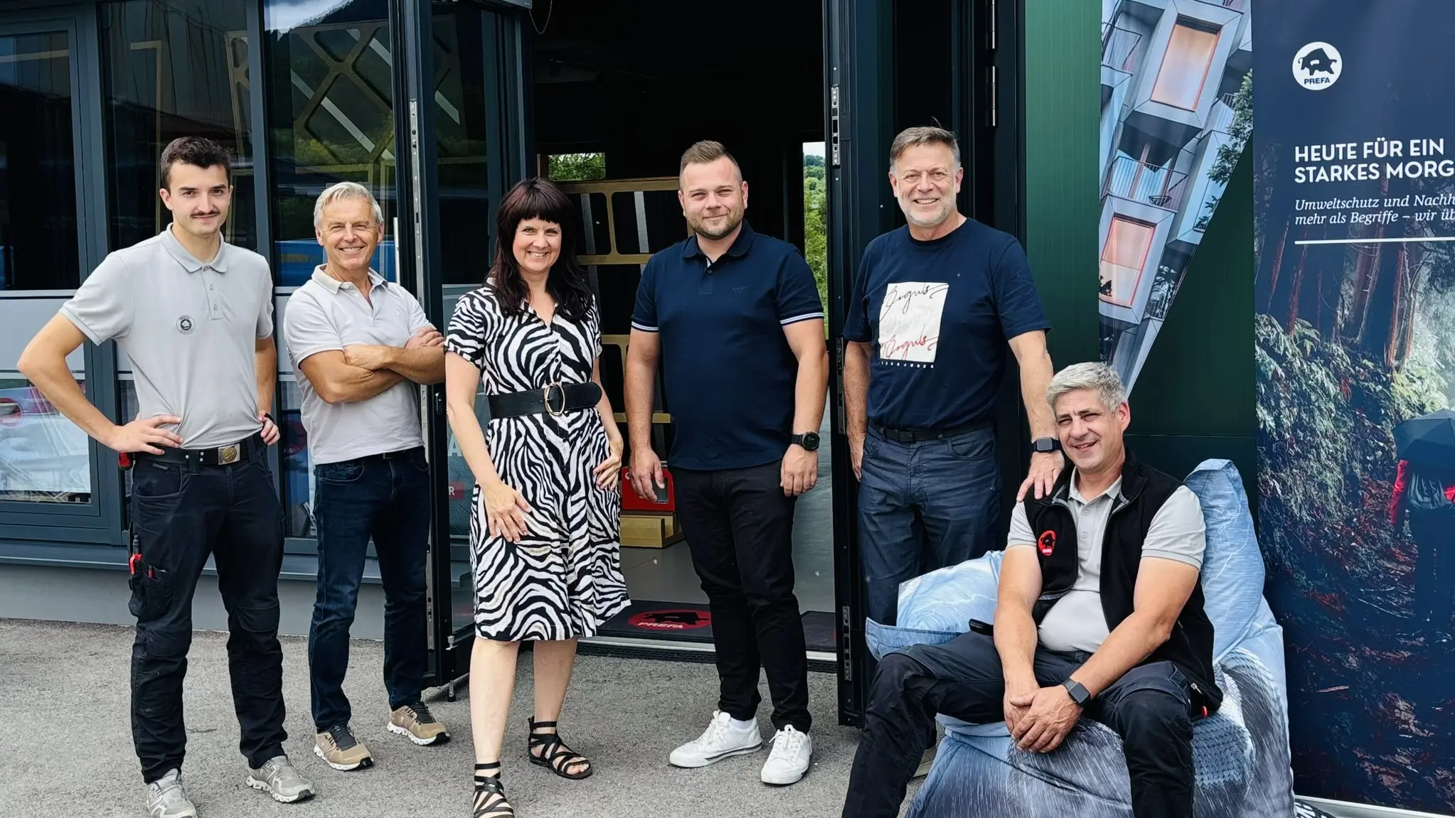 Gruppenfoto vor dem PREFA Showroom in St. Veit an der Glan: Mehrere PREFA Mitarbeiter stehen und sitzen gemeinsam vor dem Eingang. Neben der geöffneten Tür ist ein PREFA Banner mit Architekturprojekten und Aluminium-Dach- und Fassadenlösungen sichtbar. Die moderne Gebäudefassade und der Eingangsbereich unterstreichen den professionellen Rahmen. Die Szene zeigt den persönlichen Austausch, das Netzwerk sowie die gelungene Veranstaltung rund um PREFA Aluminiumprodukte für Dach und Fassade.