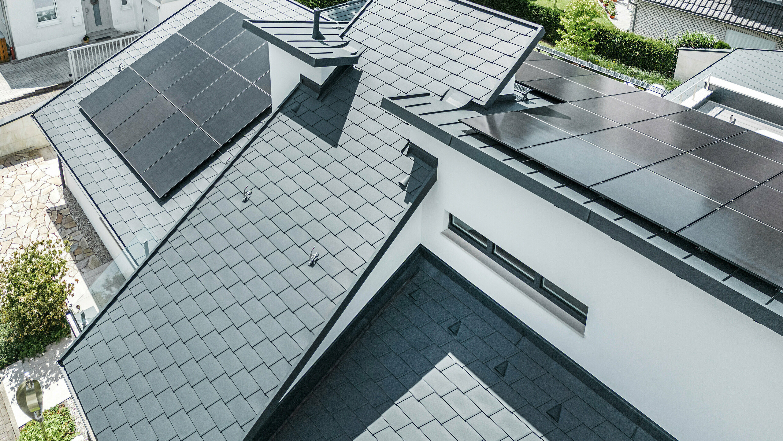 Modernes Einfamilienhaus in Dortmund mit einer fortschrittlichen Photovoltaikanlage, installiert auf einem PREFA Dachsystem bestehend aus Dachschindeln DS.19 und PREFALZ Stehfalzdeckung in der Farbe P.10 Anthrazit. Die PV-Anlage ist mittels Vario Solarhalter für die Dachschindeln und des PREFALZ Vario Solarhalter für die Stehfalzbahnen befestigt. Die Solaranlage gewährleistet eine effiziente Energieerzeugung aus erneuerbaren Energiequellen. Das durchdachte Dachdesign bietet eine optimale Balance zwischen Ästhetik und Funktionalität.