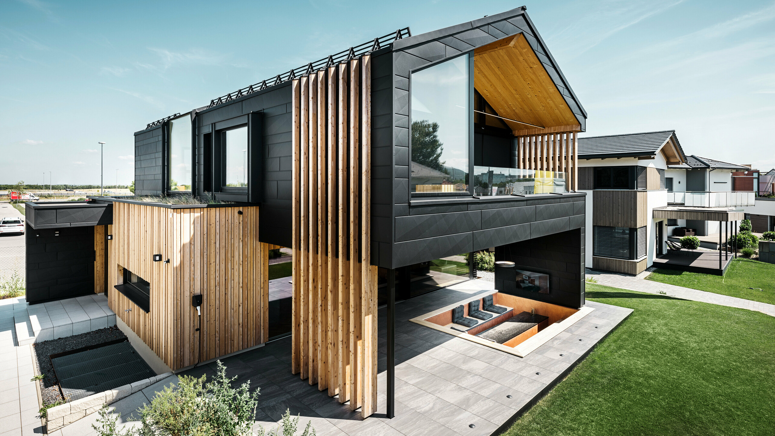Seitenansicht des Musterhauses Autark in der Blauen Lagune in Vösendorf, Österreich. Das Obergeschoss besticht durch große Fensterflächen und eine moderne Aluminiumfassade aus PREFA Siding.X Paneelen in P.10 Schwarz, kombiniert mit vertikalen Holzelementen. Die großzügige Terrasse im Erdgeschoss mit eingelassener Sitzgelegenheit fügt sich nahtlos in die Architektur ein. Das stilvolle und nachhaltige Design des Hauses verbindet Aluminium und Holz auf eindrucksvolle Art und Weise.