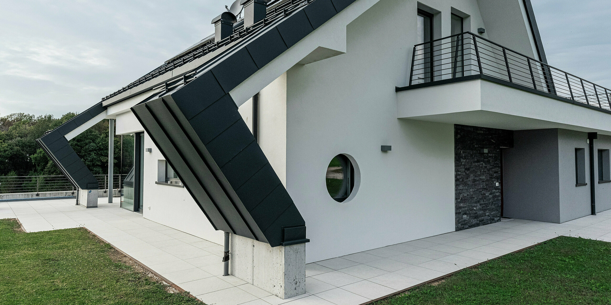 Nahaufnahme der Satteldacherweiterung eines modernen Einfamilienhauses in Borgo Valbelluna mit einer PREFALZ Stehfalzdeckung in P.10 Anthrazit. Der markante Dachüberstand des Aluminiumdaches unterstreicht die innovative Architektur und schützt die Fassade. Die langlebige, witterungsbeständige Dacheindeckung harmoniert perfekt mit der weißen Putzfassade und den dunklen Akzenten. Ein umlaufender Balkon und runde Fensterdetails verleihen dem Gebäude eine besondere Note. Hochwertiges PREFA Aluminium sorgt für Stabilität, Eleganz und Wartungsfreiheit.