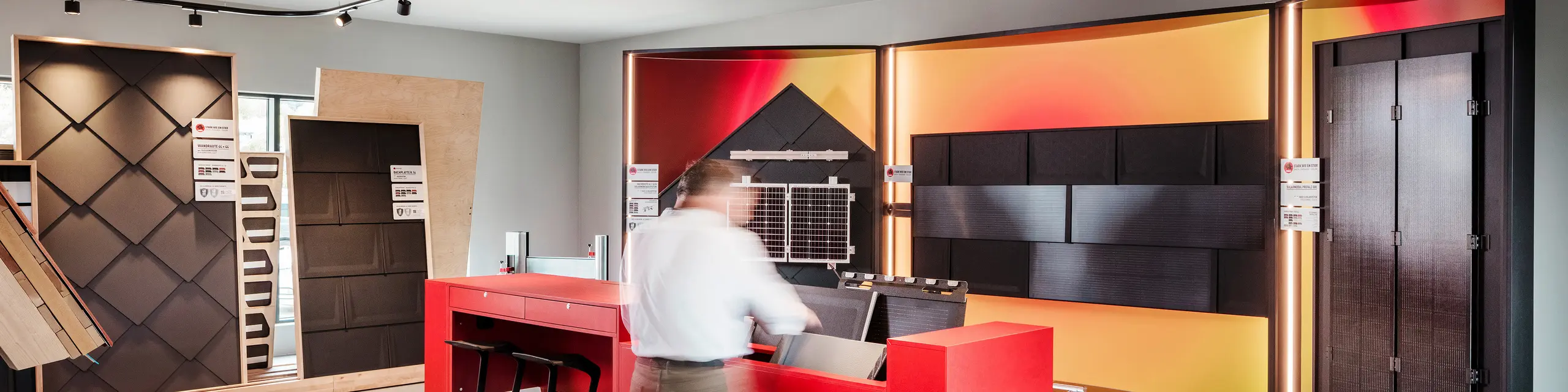 Moderner Showroom mit Ausstellung von verschiedenen PREFA Dach- und Fassadensystemen, inklusive Solardachplatten. Ein roter Tisch mit Prospektmaterial steht im Vordergrund, während verschiedene Dachmuster an den Wänden präsentiert werden.