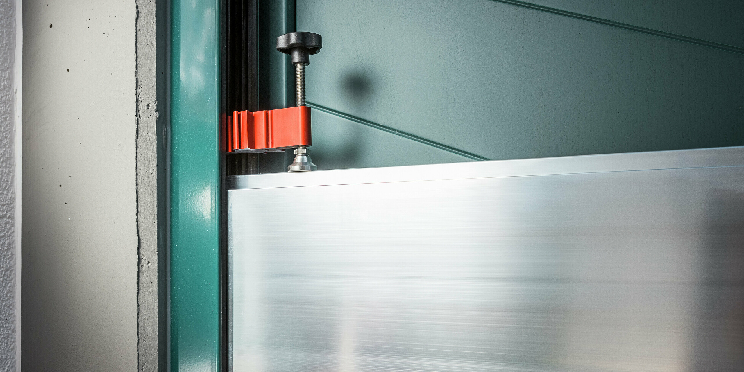 Nahaufnahme der Spannvorrichtung des PREFA Hochwasserschutzsystems im Türbereich eines Einfamilienhauses in Schwaz. Zu sehen ist die präzise Fixierung der Aluminium Dammbalken in den seitlichen Wandprofilen mittels rotem Spannbügel und schwarzer Schraubklemme. Die hochwertige Verarbeitung und die einfache Handhabung unterstreichen die Alltagstauglichkeit des Systems - ideal für den schnellen Hochwasserschutz an Hauseingängen.