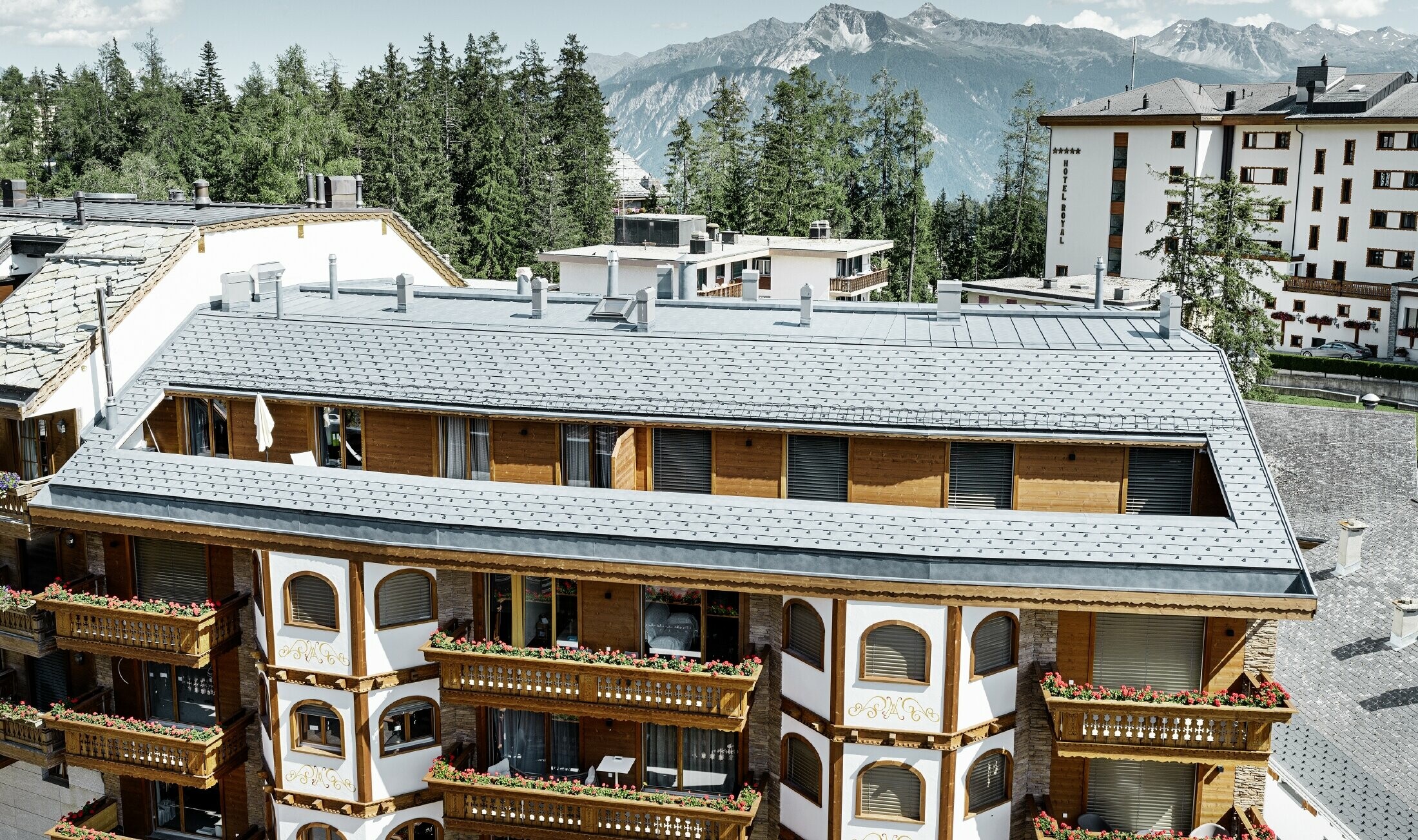 Mehrfamilienhaus in Crans-Montana mit Bergen im Hintergrund und einer Fassade mit verspielten Holzelementen mit einem PREFA Aluminium Dachschindel-Dach in steingrau 