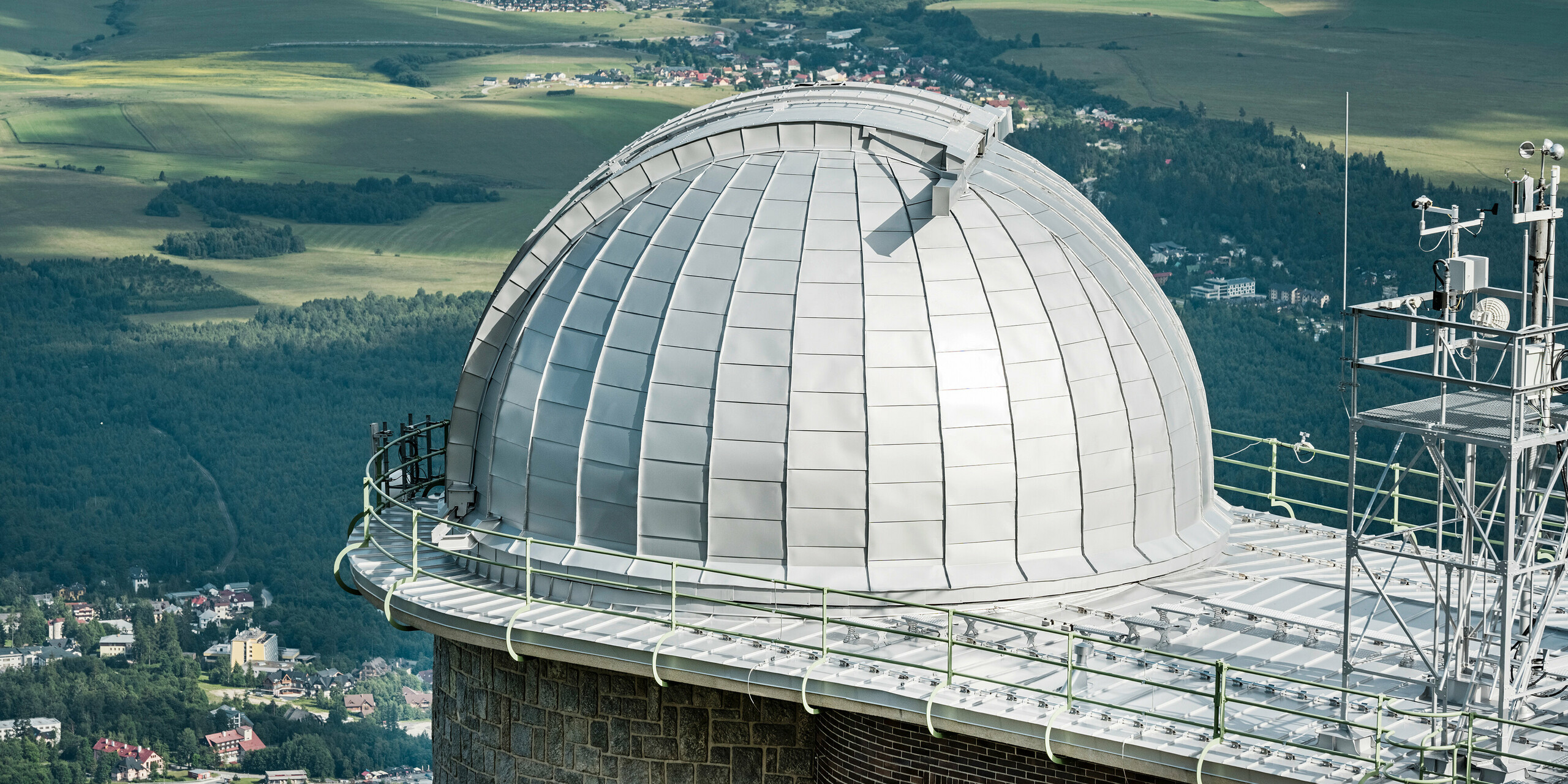 Panoramablick auf die imposante Kuppel der Sternwarte Skalnaty Pleso, meisterhaft eingebettet in die malerische Landschaft der Hohen Tatra. Die silbermetallic PREFALZ Aluminiumverkleidung schimmert im Kontrast zum üppigen Grün der umliegenden Wälder und den sanften Konturen der ländlichen Landschaft. Dieses architektonische Juwel, das auf einer Höhe von 1.783 Metern über dem Meeresspiegel ruht, ist nicht nur ein Wahrzeichen für astronomische Entdeckungen seit 1943, sondern auch ein Zeugnis für die Langlebigkeit und Umweltverträglichkeit von PREFA Aluminiumprodukten, die durch eine 40-jährige Garantie untermauert sind.