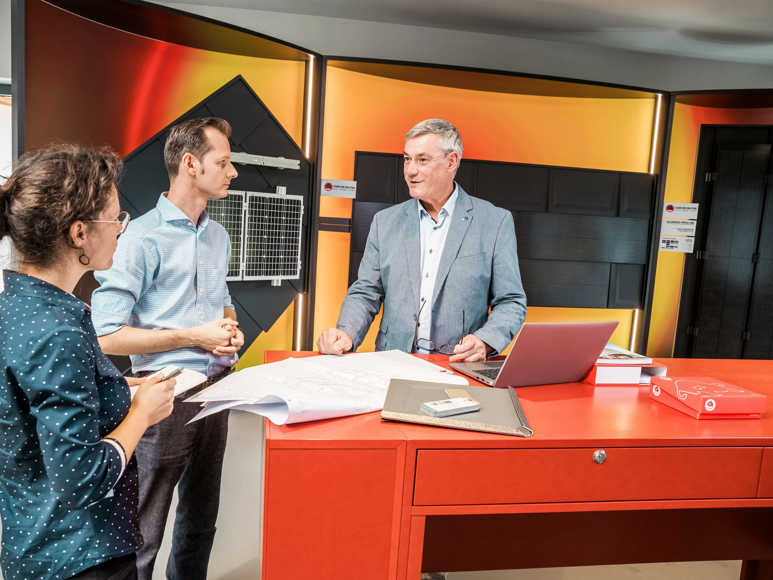 Ein Architekt und ein Berater diskutieren über Baupläne in einem modernen Showroom, der mit PREFA Dach- und Fassadenlösungen ausgestattet ist. Im Hintergrund sind verschiedene Solardach- und Fassadenplatten zu sehen.